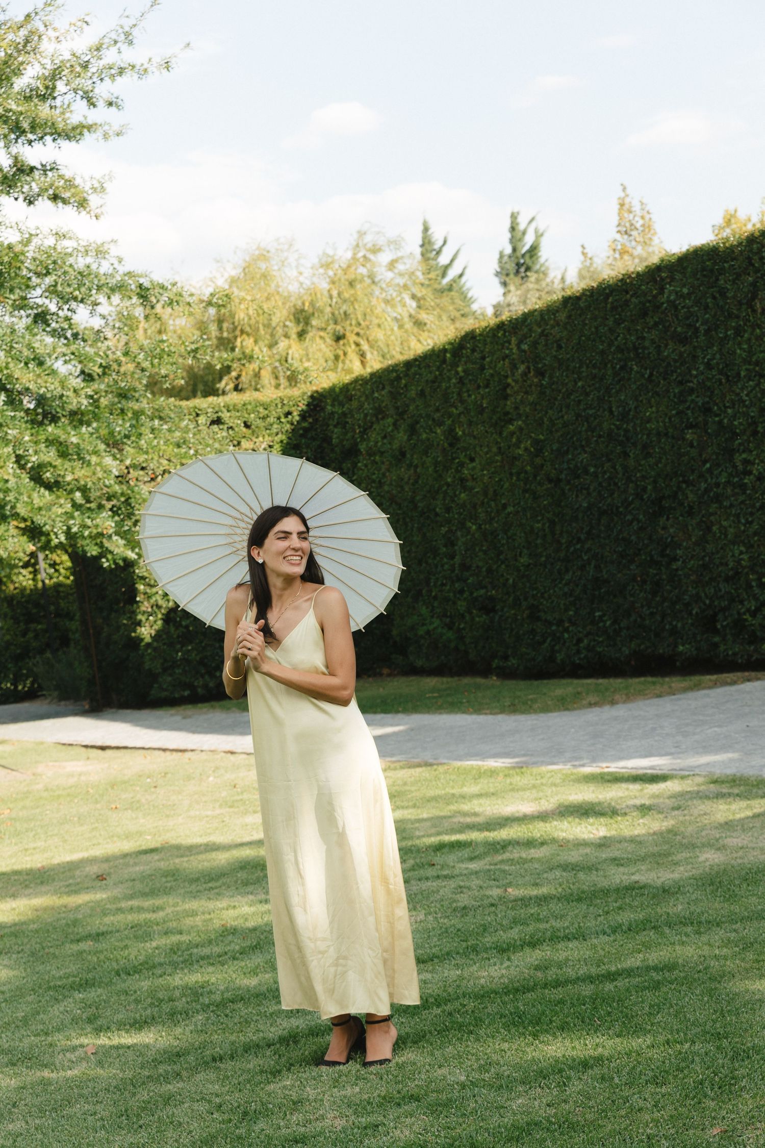 Figure with parasol poses playfully on manicured lawn with hedge backdrop.
