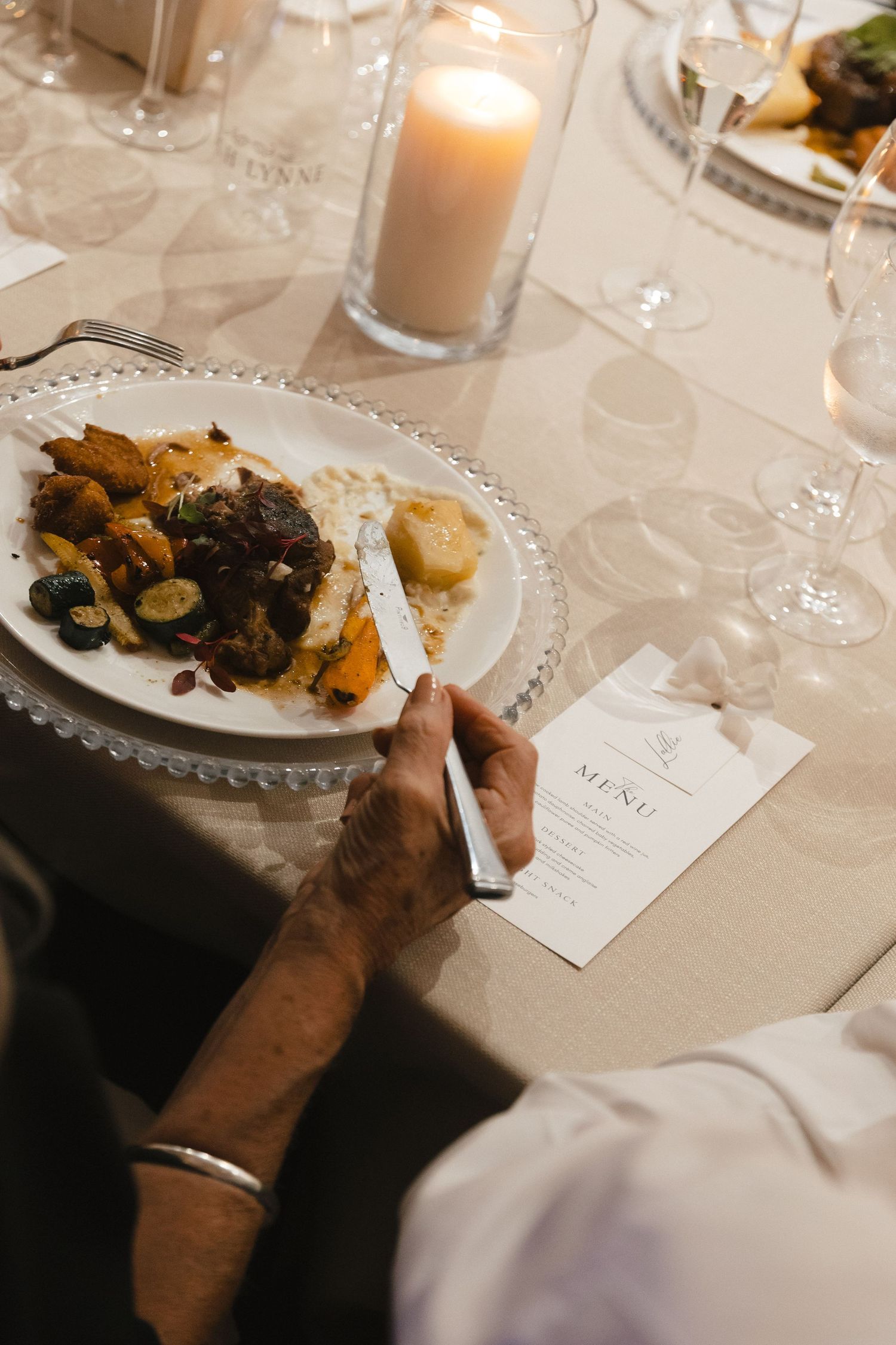 Elegant wedding reception table settings with candles, menus and plated meals in warm lighting.