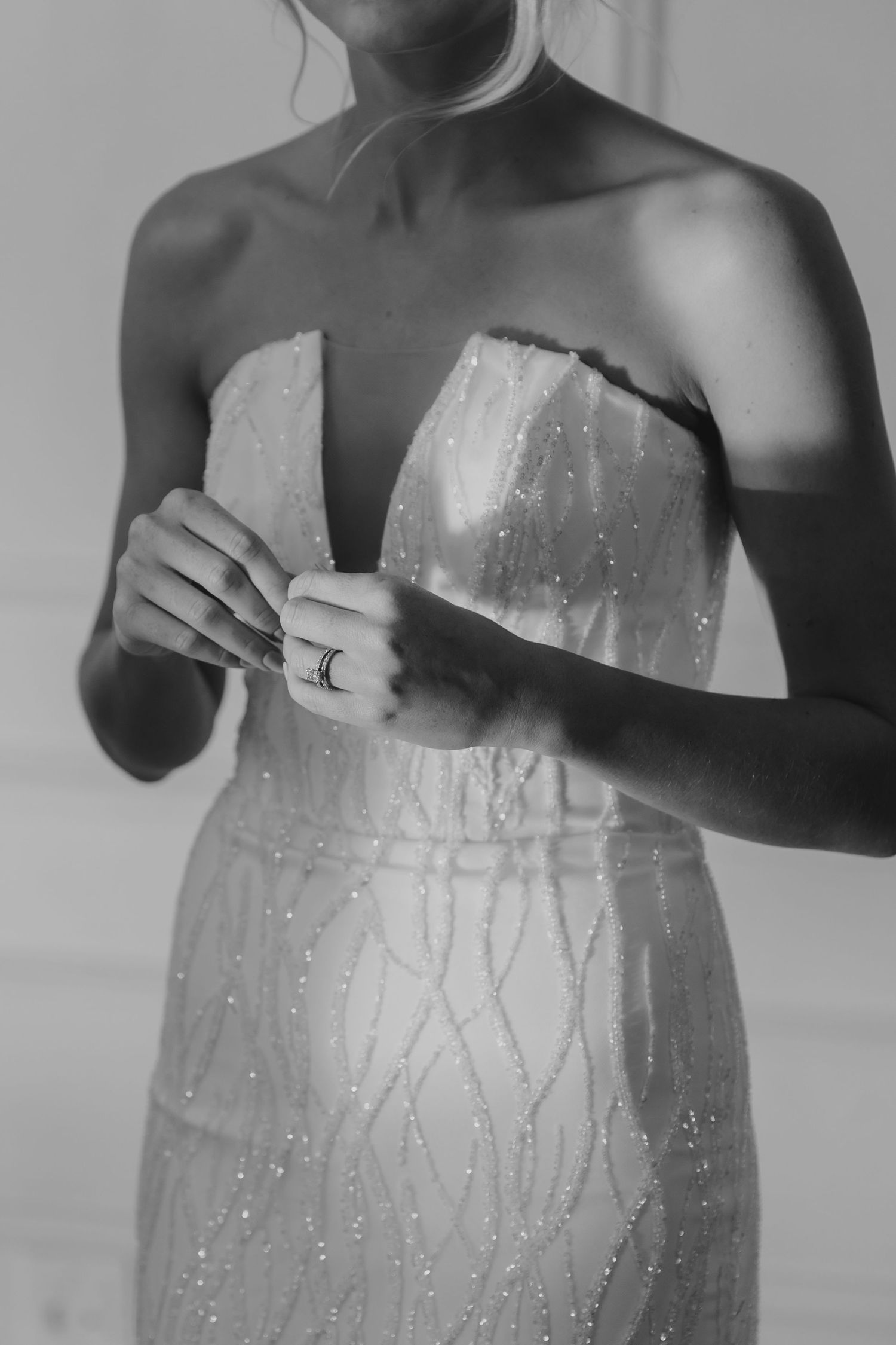 Black and white photo of a bride in a strapless beaded wedding gown, adjusting her dress with elegant rings on her fingers.