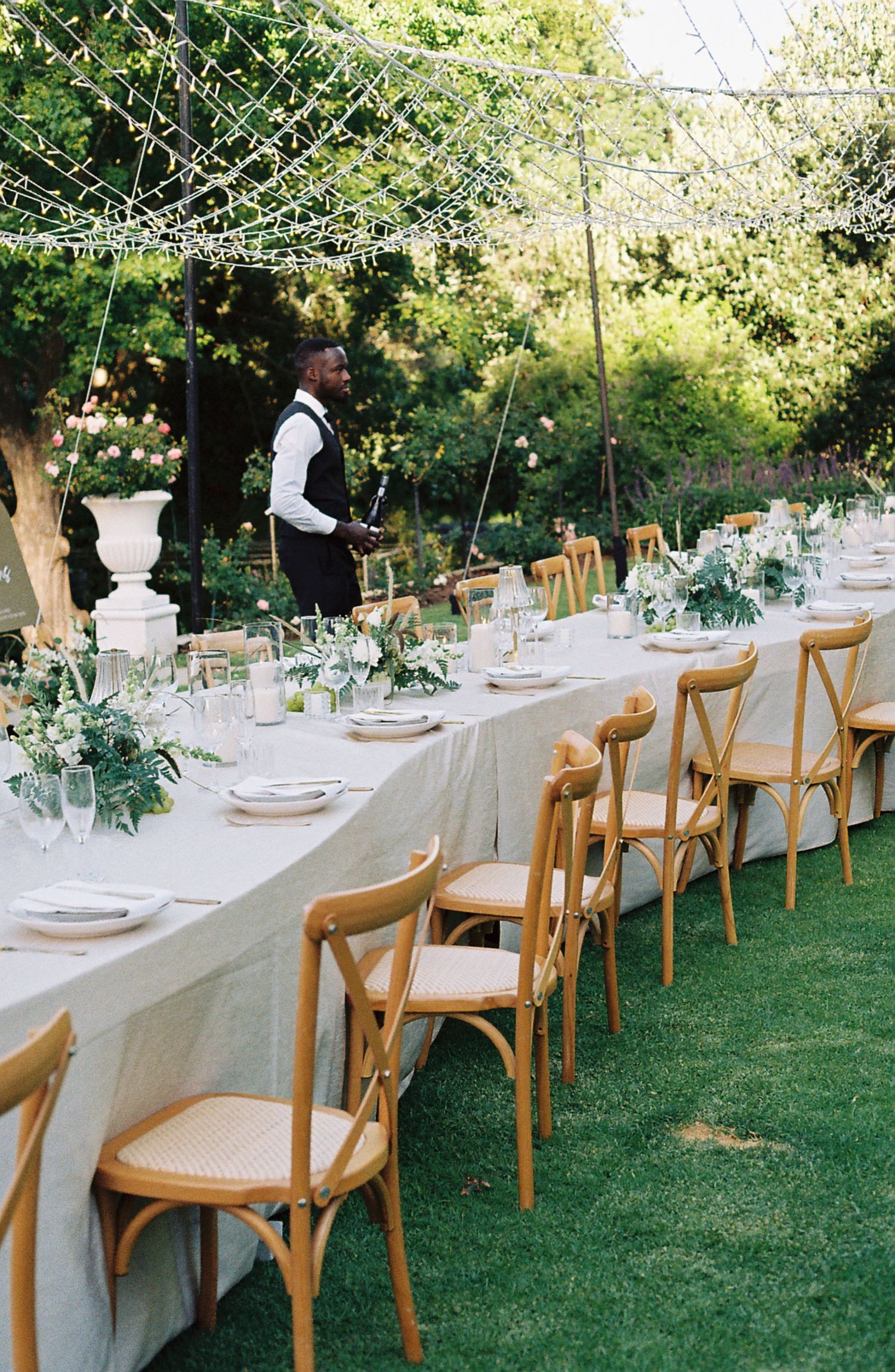 Long white dining tables with wooden chairs set up outdoors on a lawn for an elegant garden wedding reception.