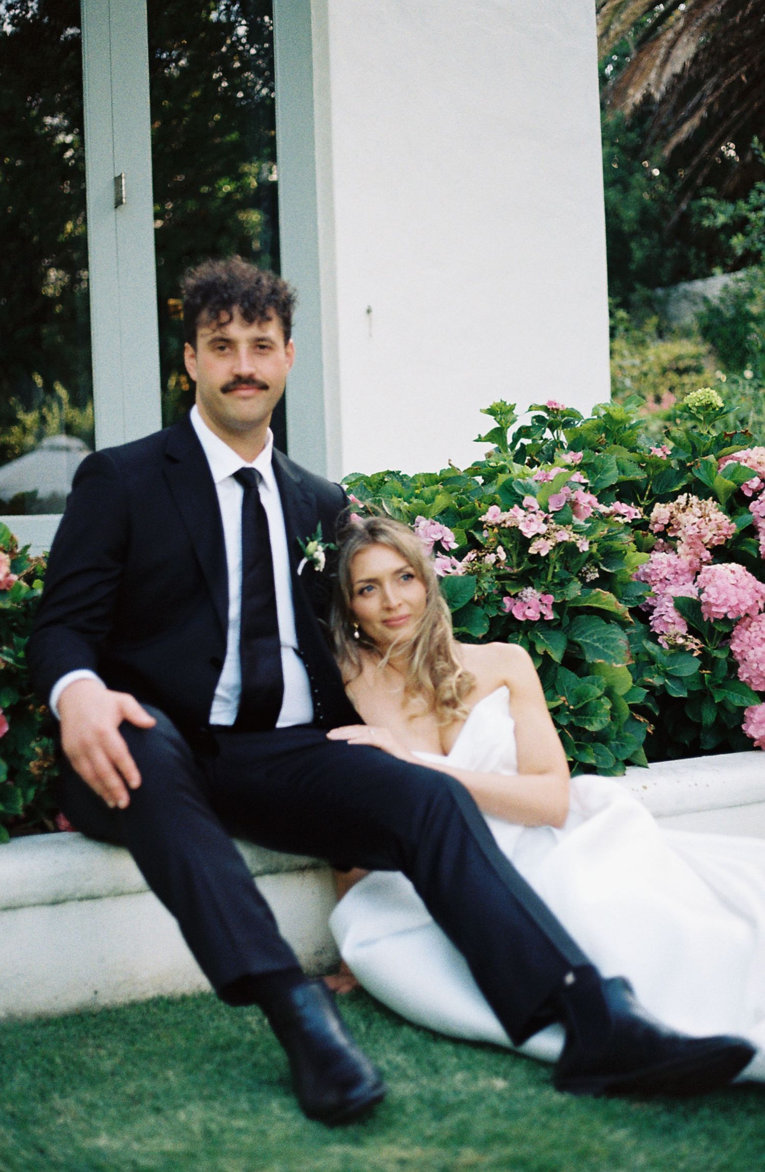 A romantic wedding couple lounges on white columns surrounded by pink hydrangeas in a garden setting.