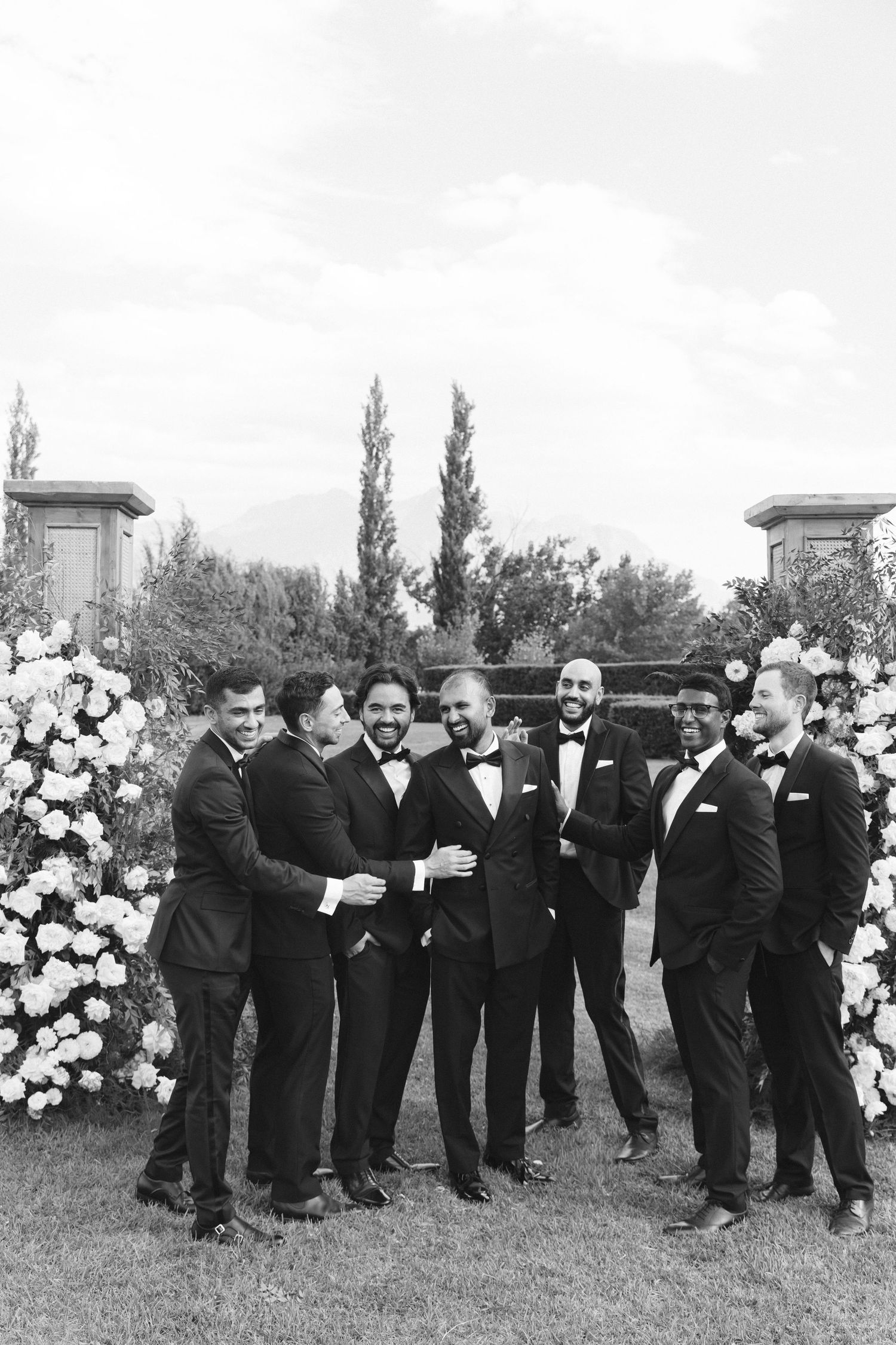 Groomsmen in black tuxedos pose together playfully on a lawn with floral arrangements visible.
