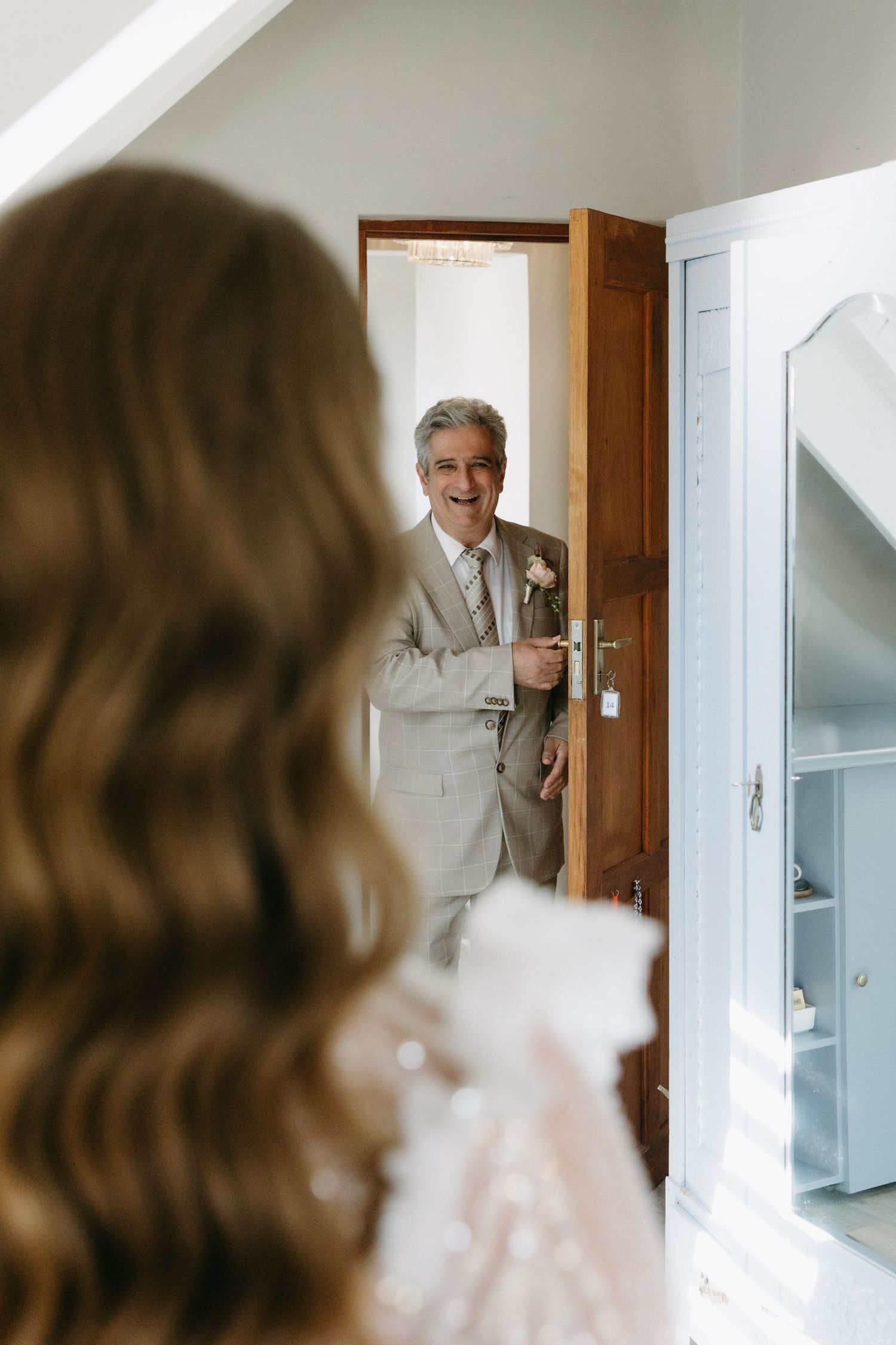 Father of the bride in a tan suit smiling at doorway during wedding morning preparations.