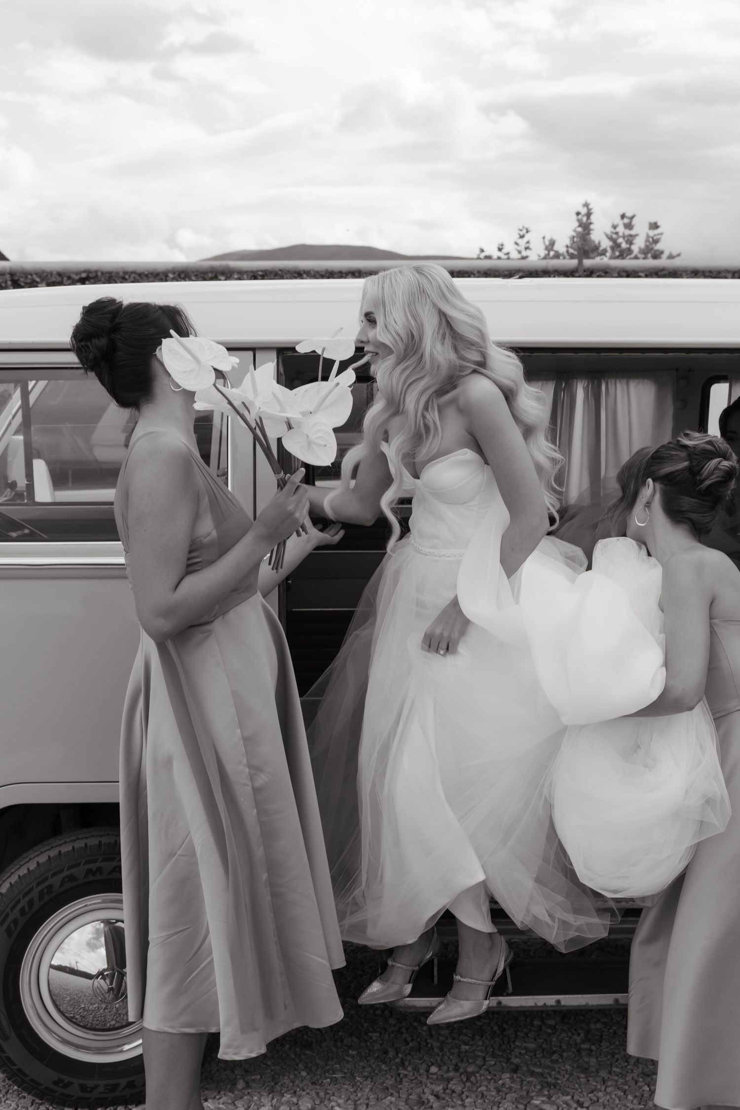 Black and white photo of bride in tulle gown with bridesmaids beside a vintage VW camper van outdoors.