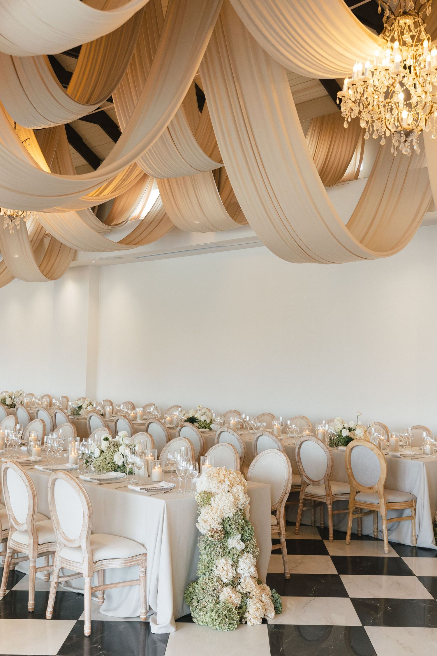 Elegant wedding reception venue with draped fabric ceiling, crystal chandeliers, and white chair covers on checkered floor.