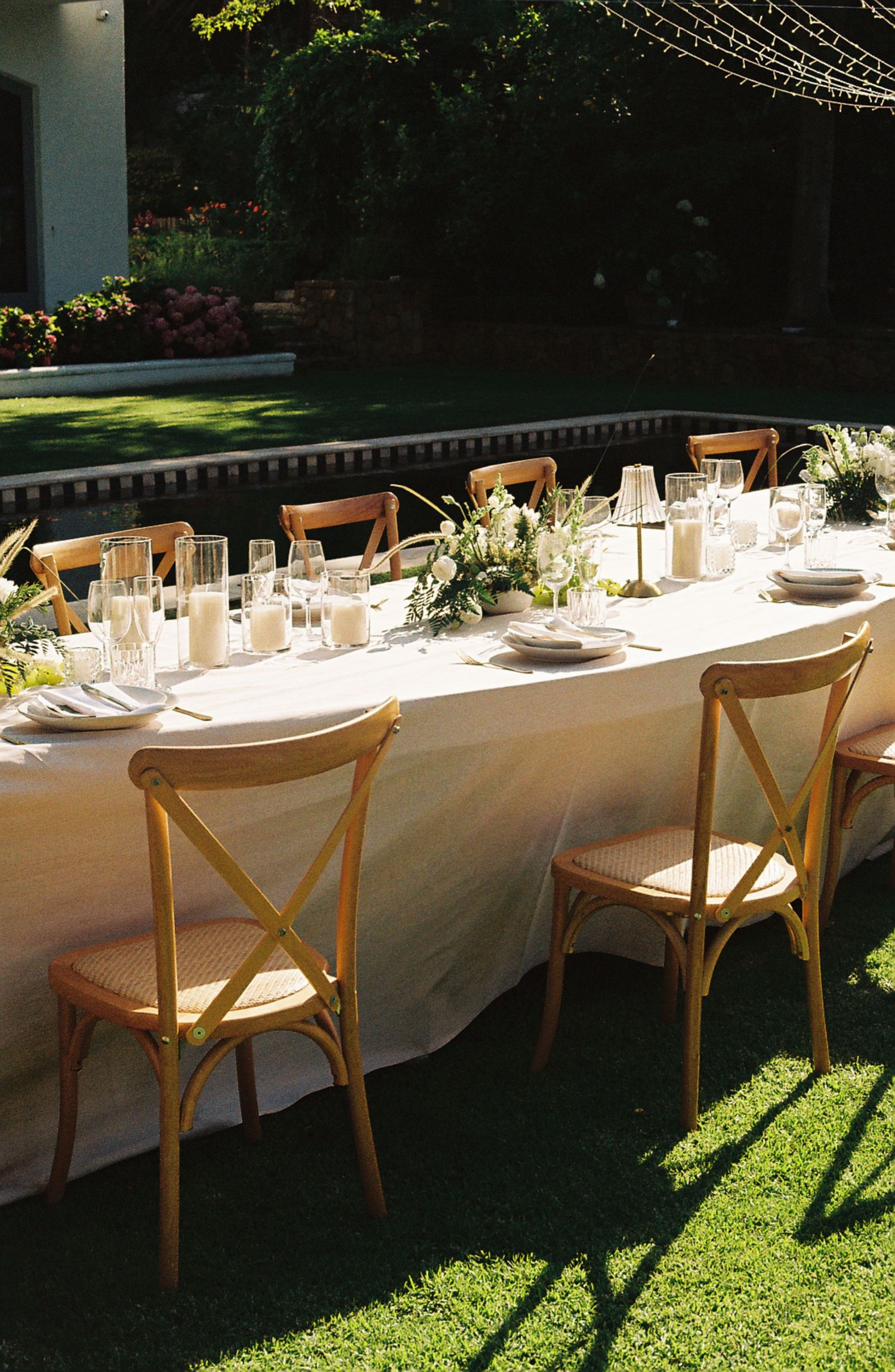 Long outdoor dining table with white linens and wooden cross-back chairs set up for an elegant garden party.