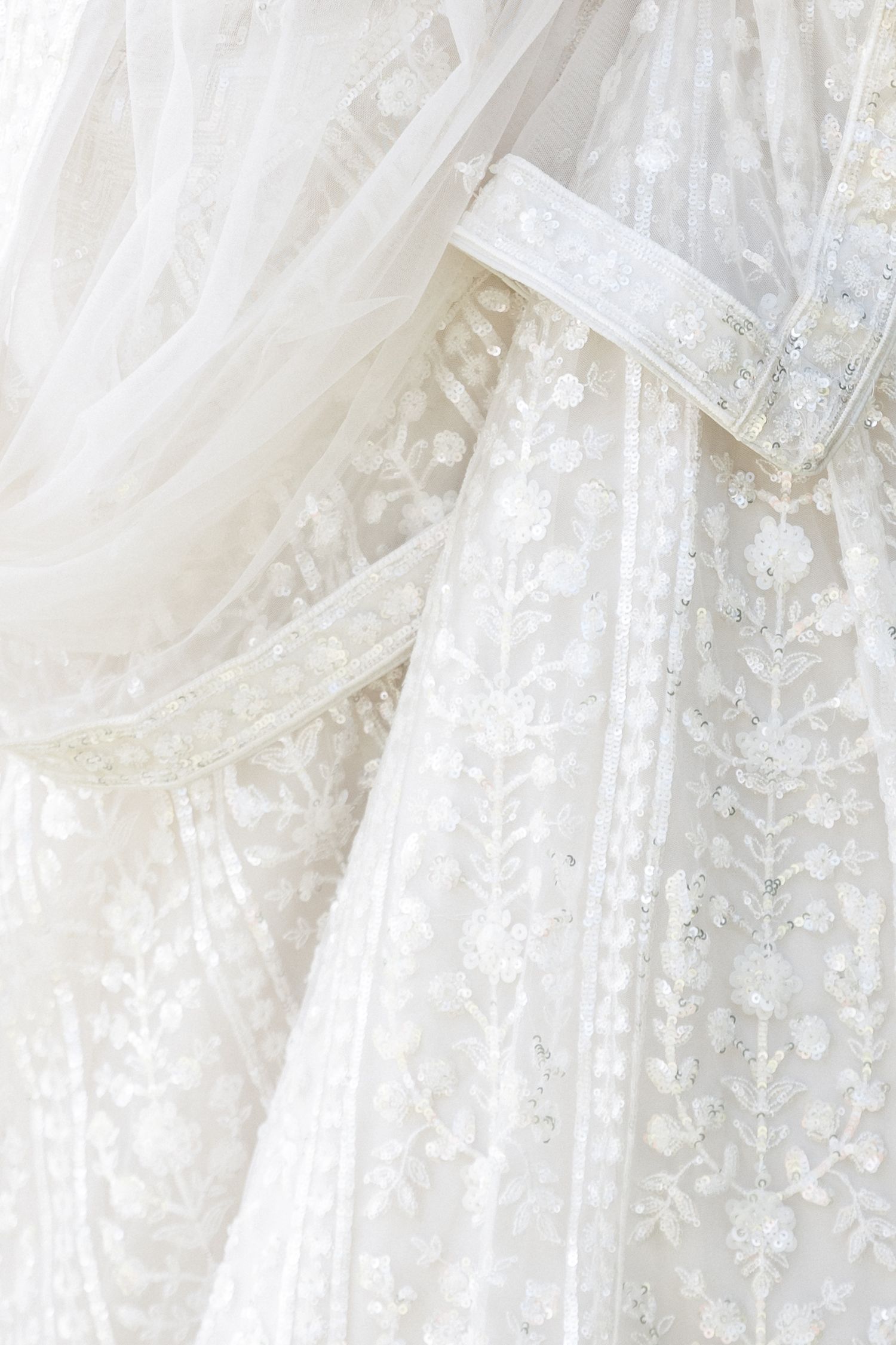 Close-up detail of delicate white lace wedding dresses with floral embroidery and beaded embellishments hanging together.