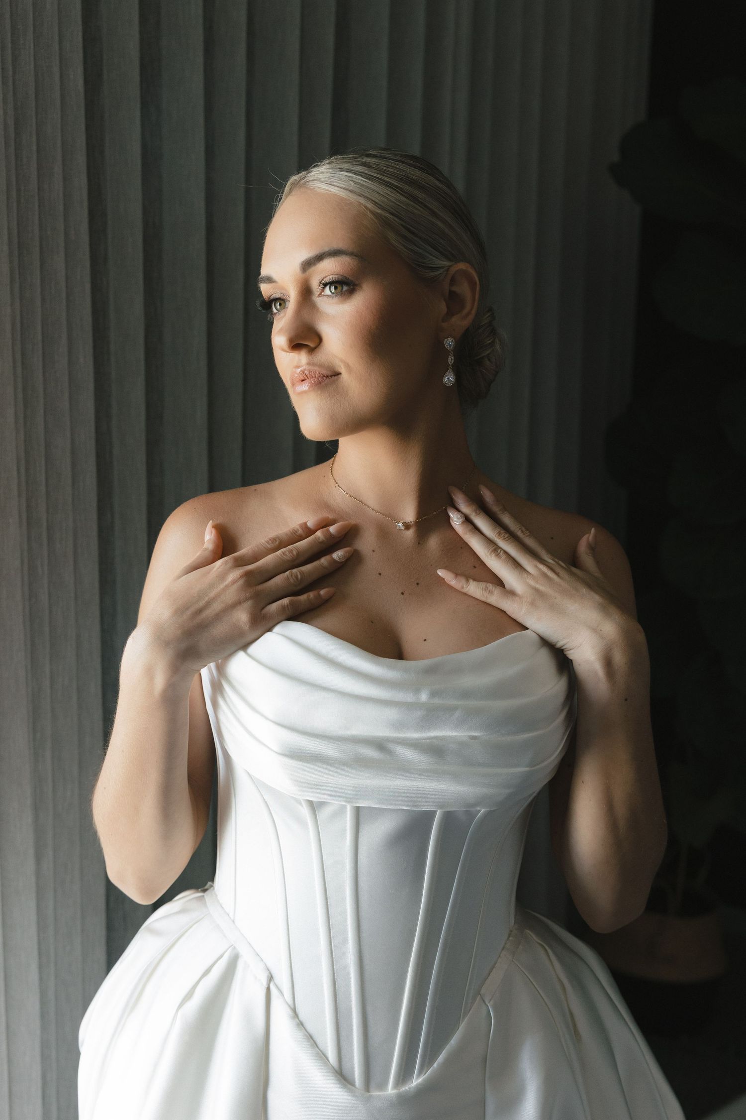 Elegant bride in white corset wedding dress poses by window in soft natural light with hands on collar.