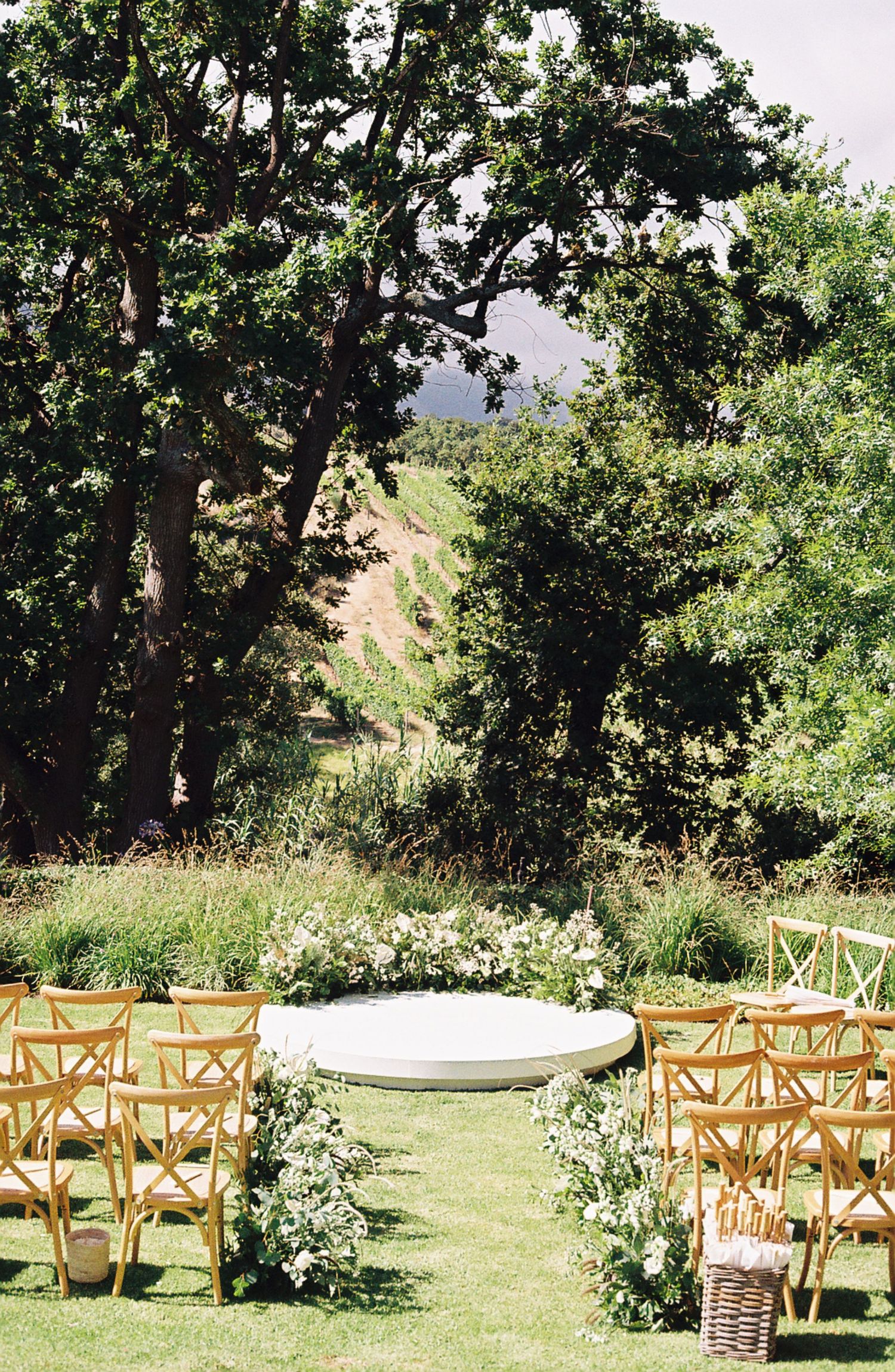 Outdoor wedding ceremony setup with wooden chairs and floral arrangements framed by lush trees in a garden setting.