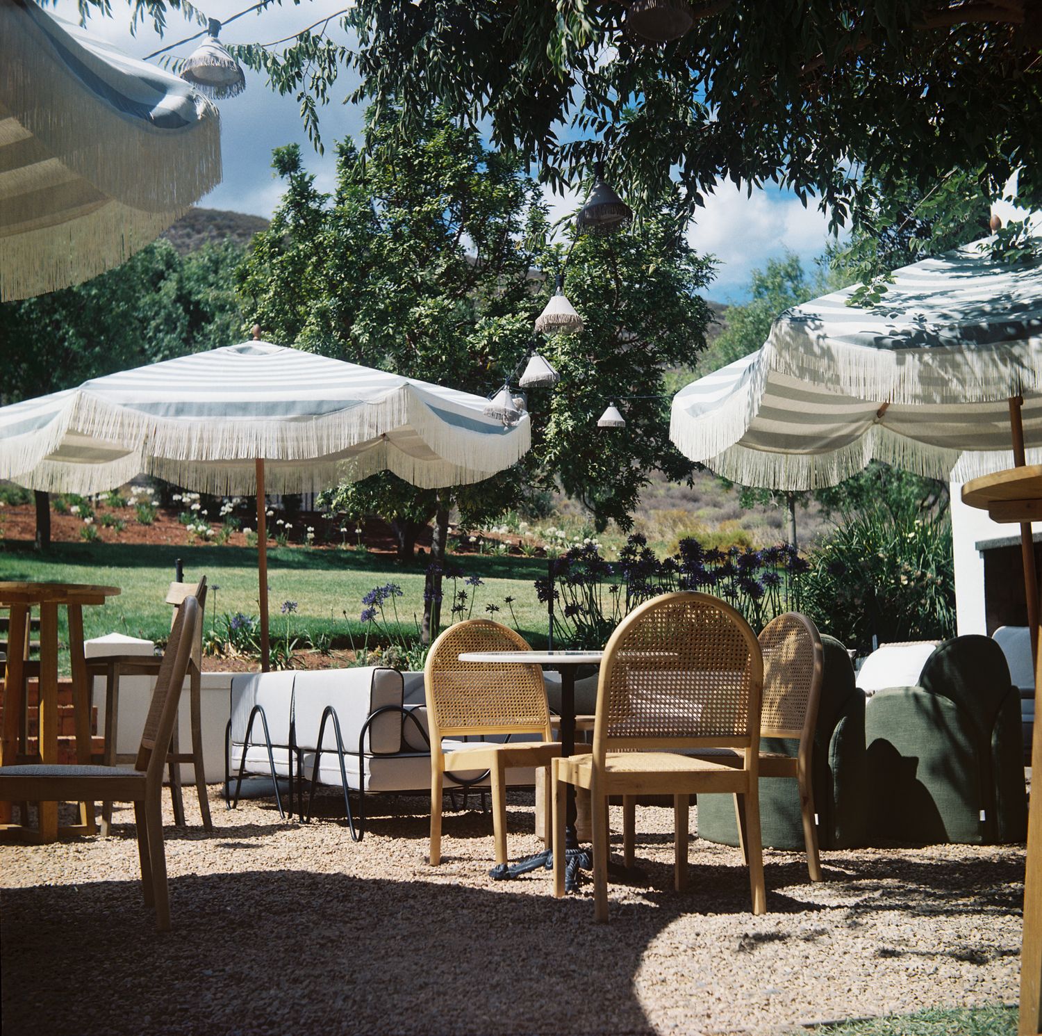 Outdoor patio seating area with white umbrellas and wicker chairs under shady trees on a sunny summer day.