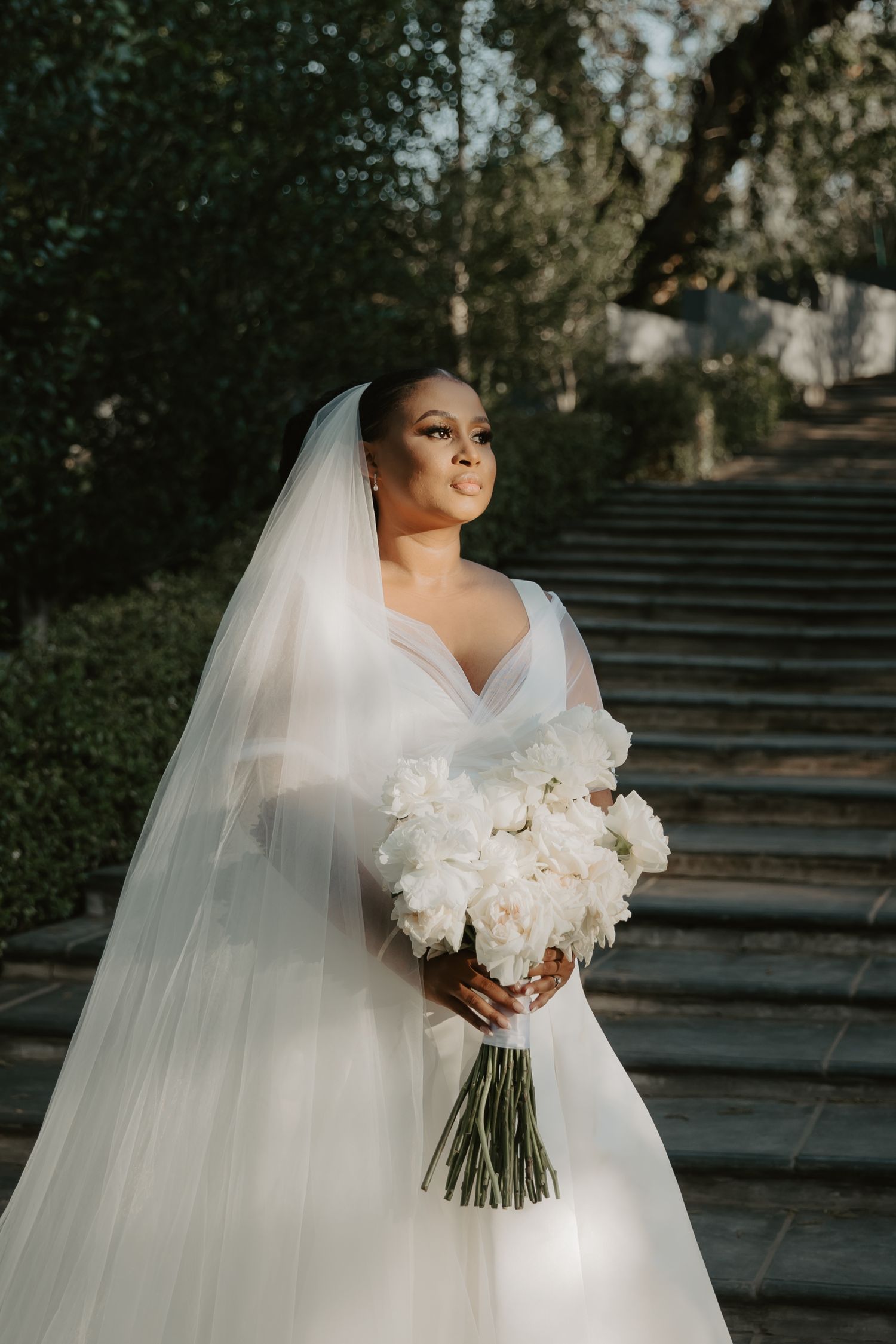 Bride in white gown and veil holds white peony bouquet on stone steps surrounded by lush greenery.