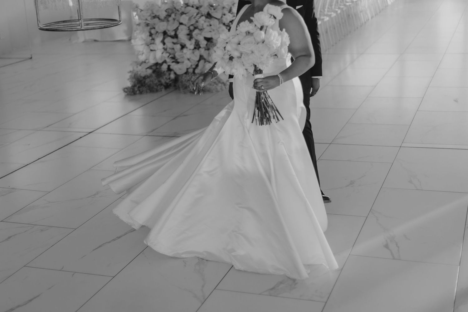 Black and white photo of a bride in a flowing white gown holding a bouquet, dancing with groom at wedding reception.