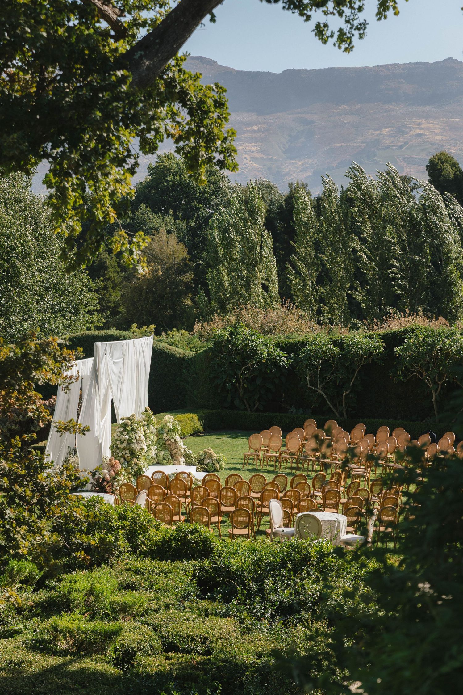 Outdoor wedding ceremony setup with wooden chairs arranged in rows facing mountains, with white fabric draping and trees.