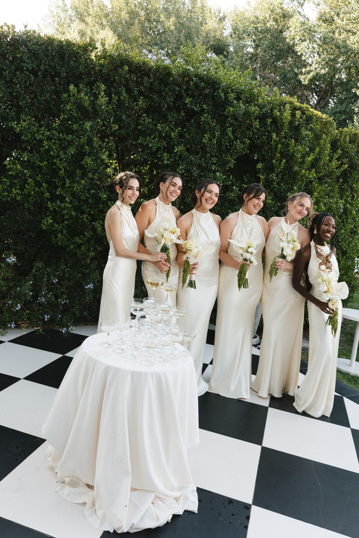 Bridesmaids in elegant cream-colored dresses pose with bouquets on a black and white checkered floor next to champagne table.