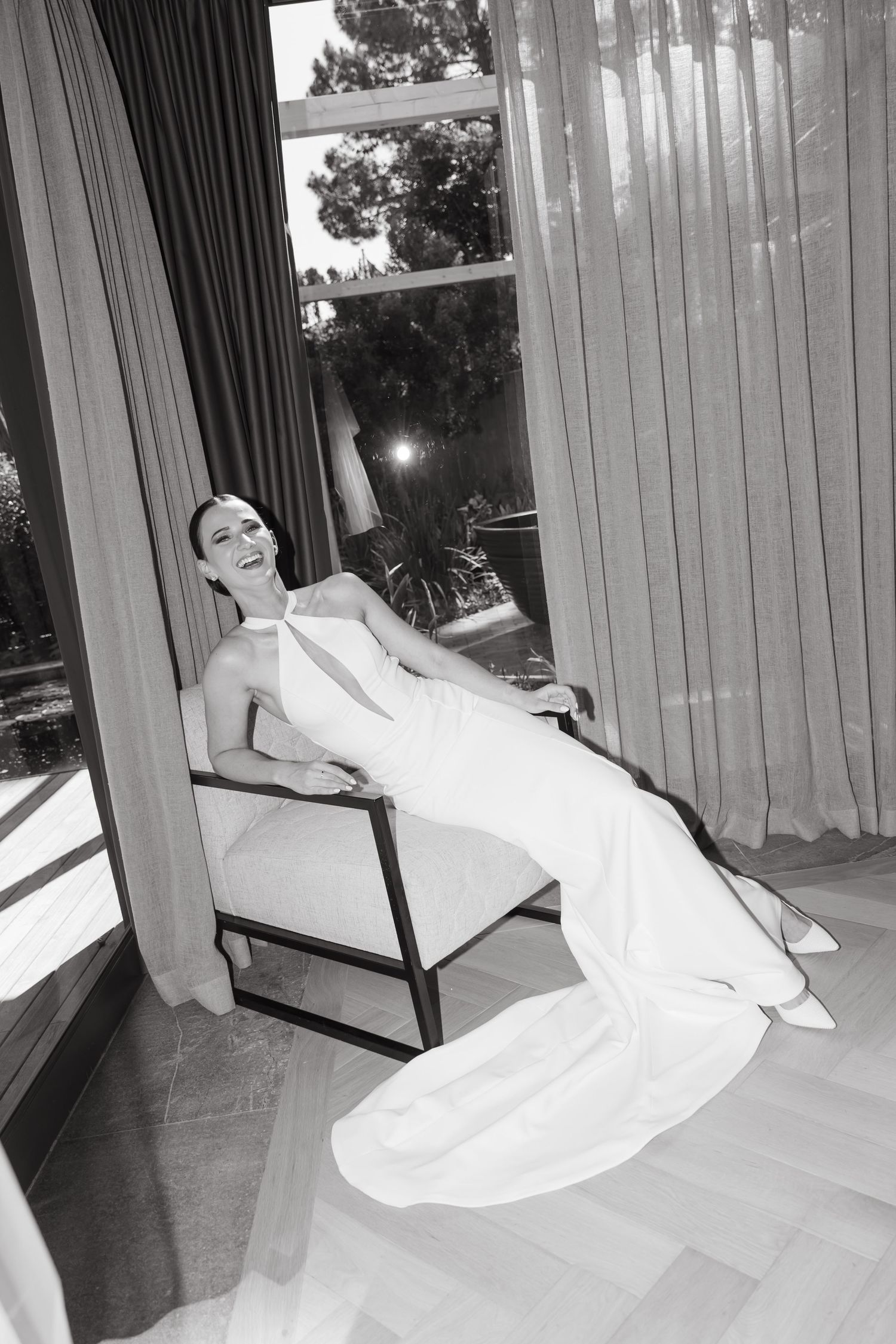 Black and white photo of a bride in a white gown laughing while seated in a chair near sheer curtains.