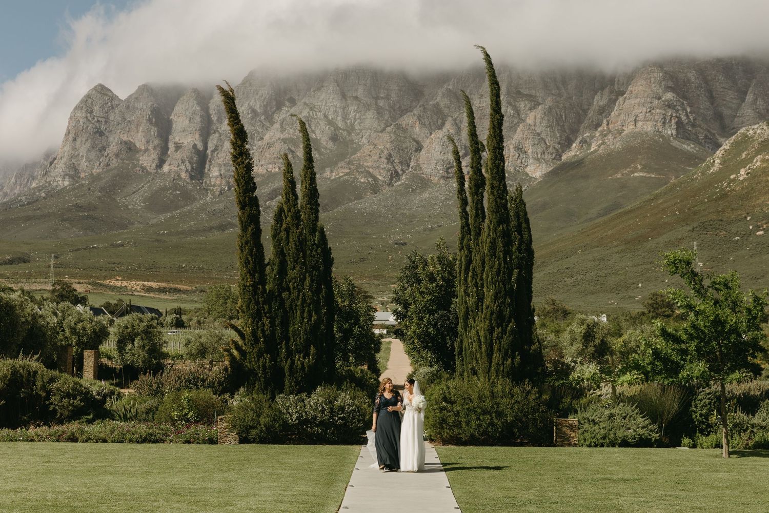 Couple walks garden path with tall cypress trees and misty mountain backdrop at elegant outdoor wedding venue.