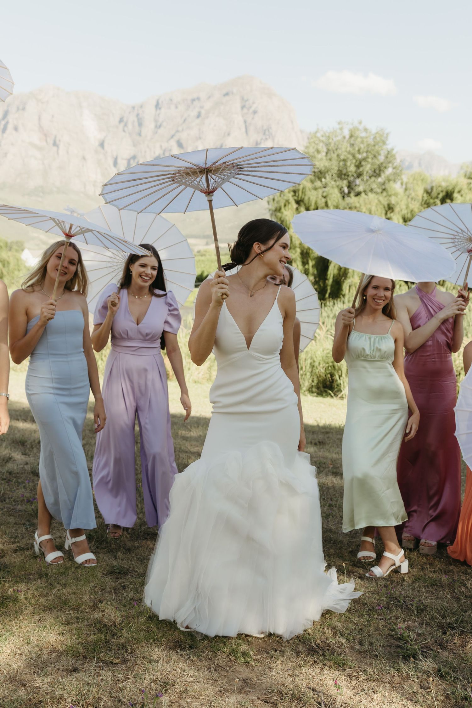 Bride in white gown with bridesmaids in colorful dresses holding white parasols outdoors with mountain backdrop.