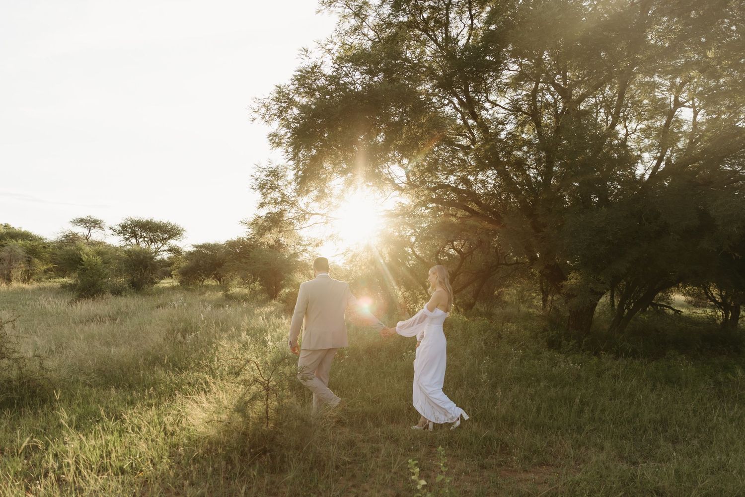 Couple holding hands dancing in golden sunlight among lush green trees in a field, woman in flowing white dress.