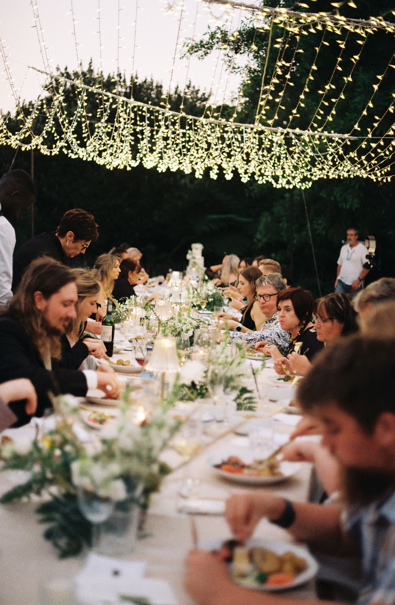 Wedding guests enjoy an outdoor dinner reception under string lights and greenery decorations at long tables.