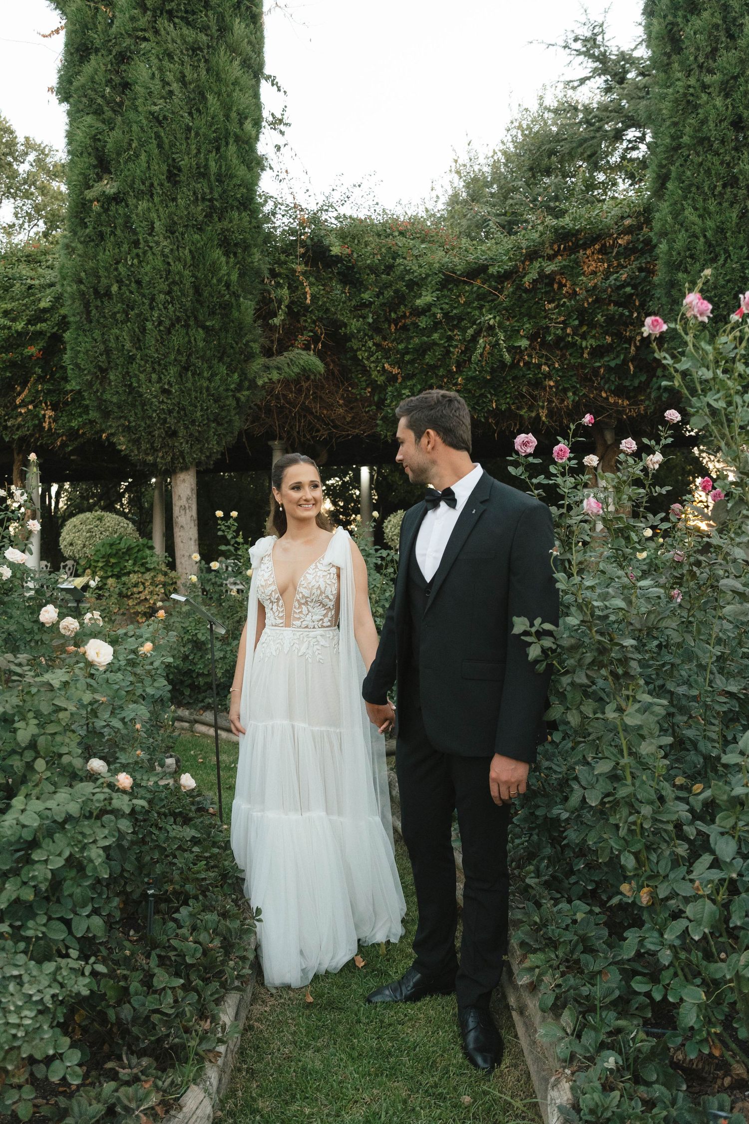 A romantic garden wedding scene with a couple standing among blooming roses and tall cypress trees.