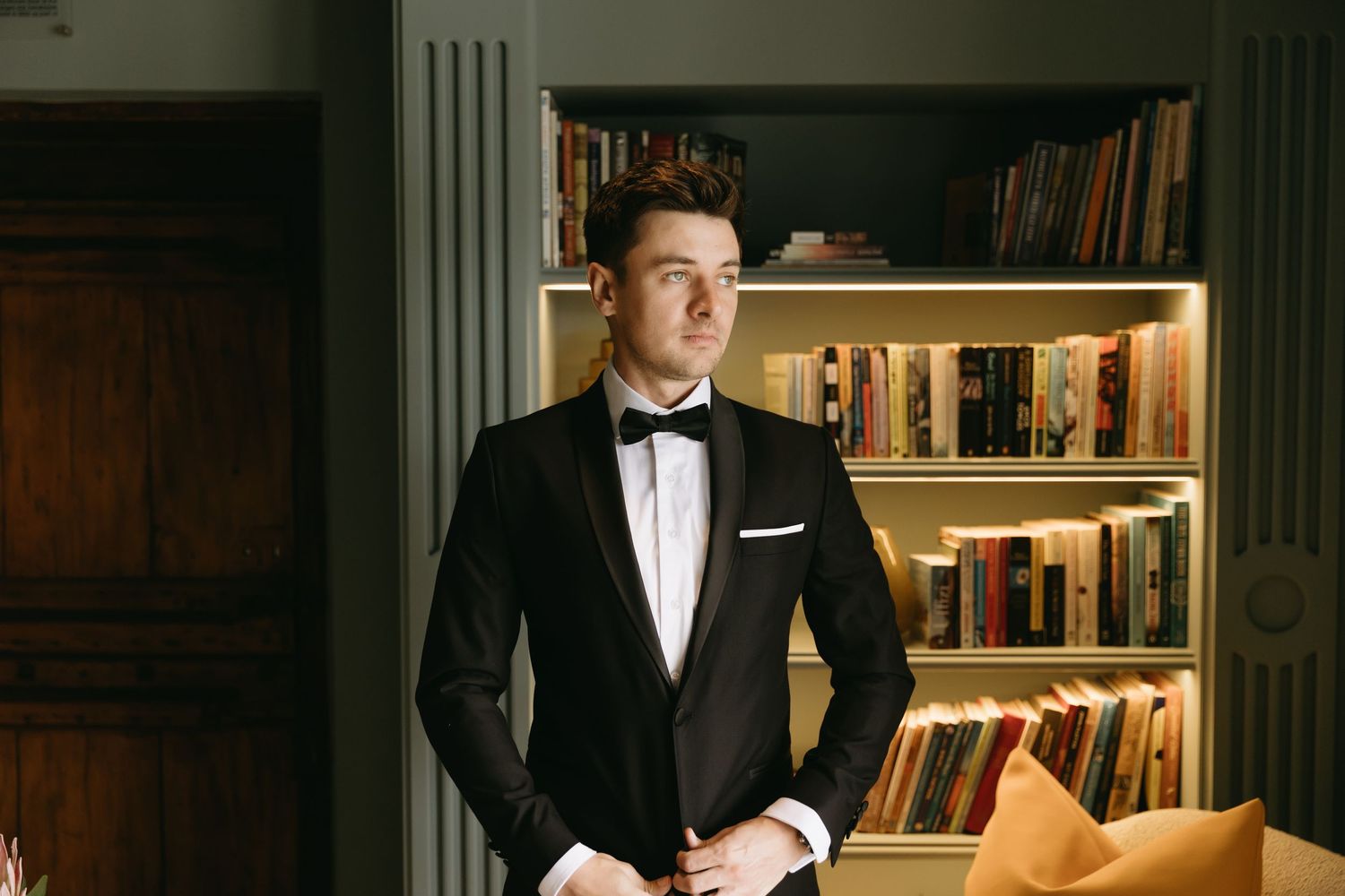 Elegant man in black tuxedo with bow tie standing before illuminated bookshelf in sophisticated interior.