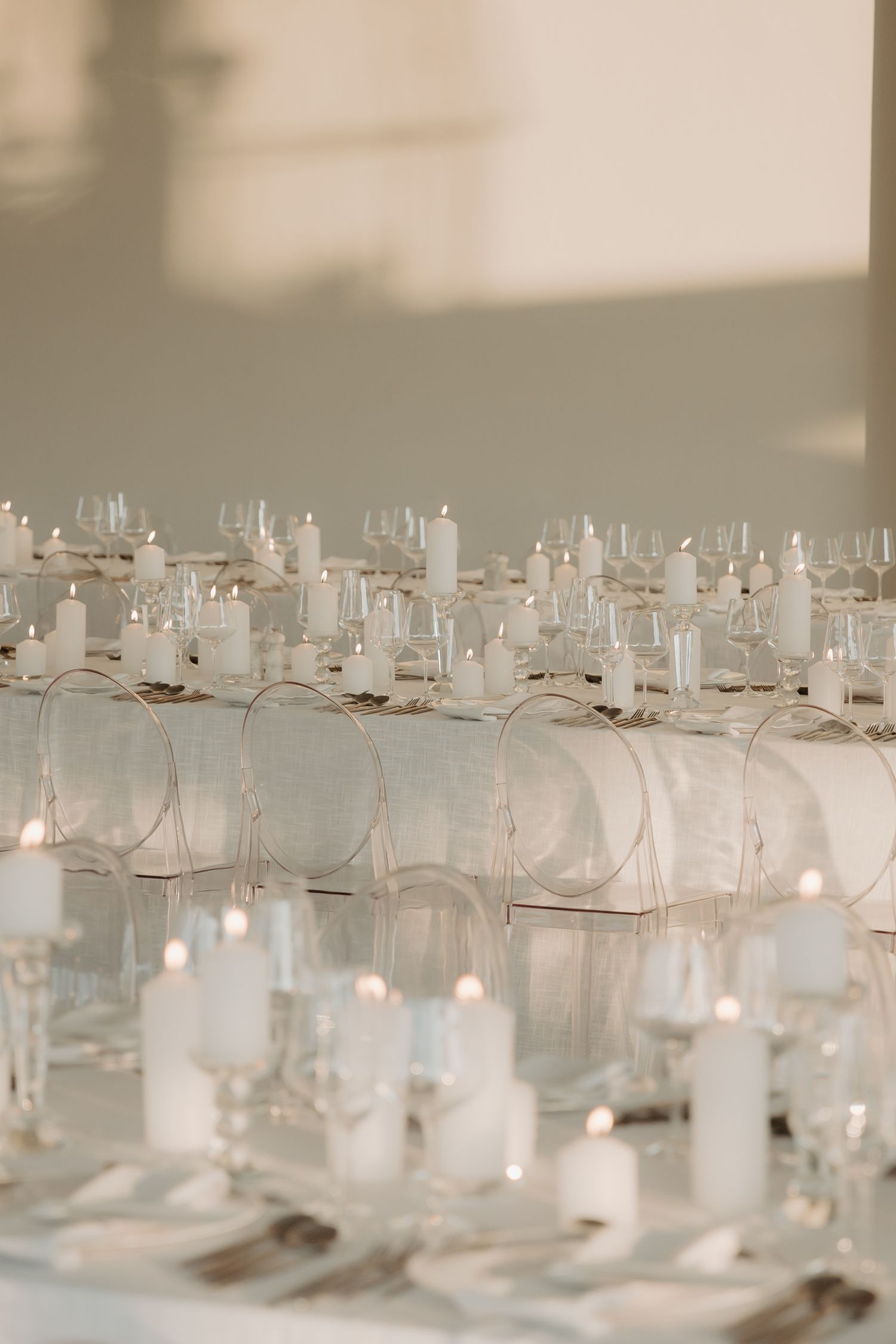 Elegant wedding reception table setup with white pillar candles, crystal glasses, and ghost chairs in a minimalist setting.