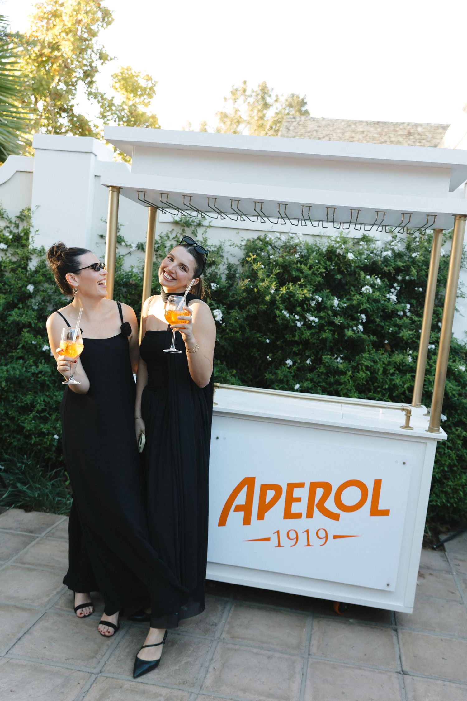Two people in black dresses enjoy drinks near a white Aperol bar cart at an outdoor event.