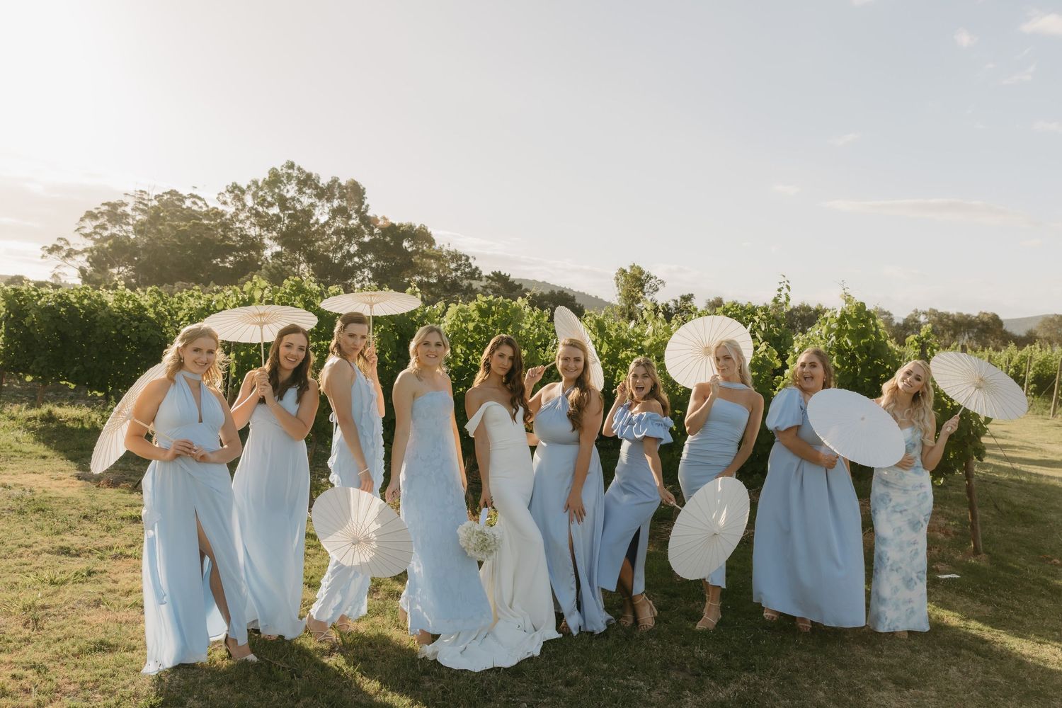 Wedding party in light blue and white dresses poses together at sunset in an outdoor garden setting.