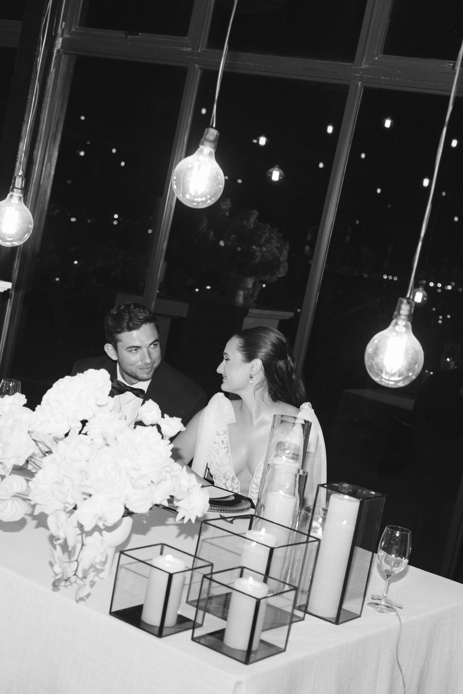 An elegant wedding reception table setting with hanging light bulbs and white floral centerpieces.