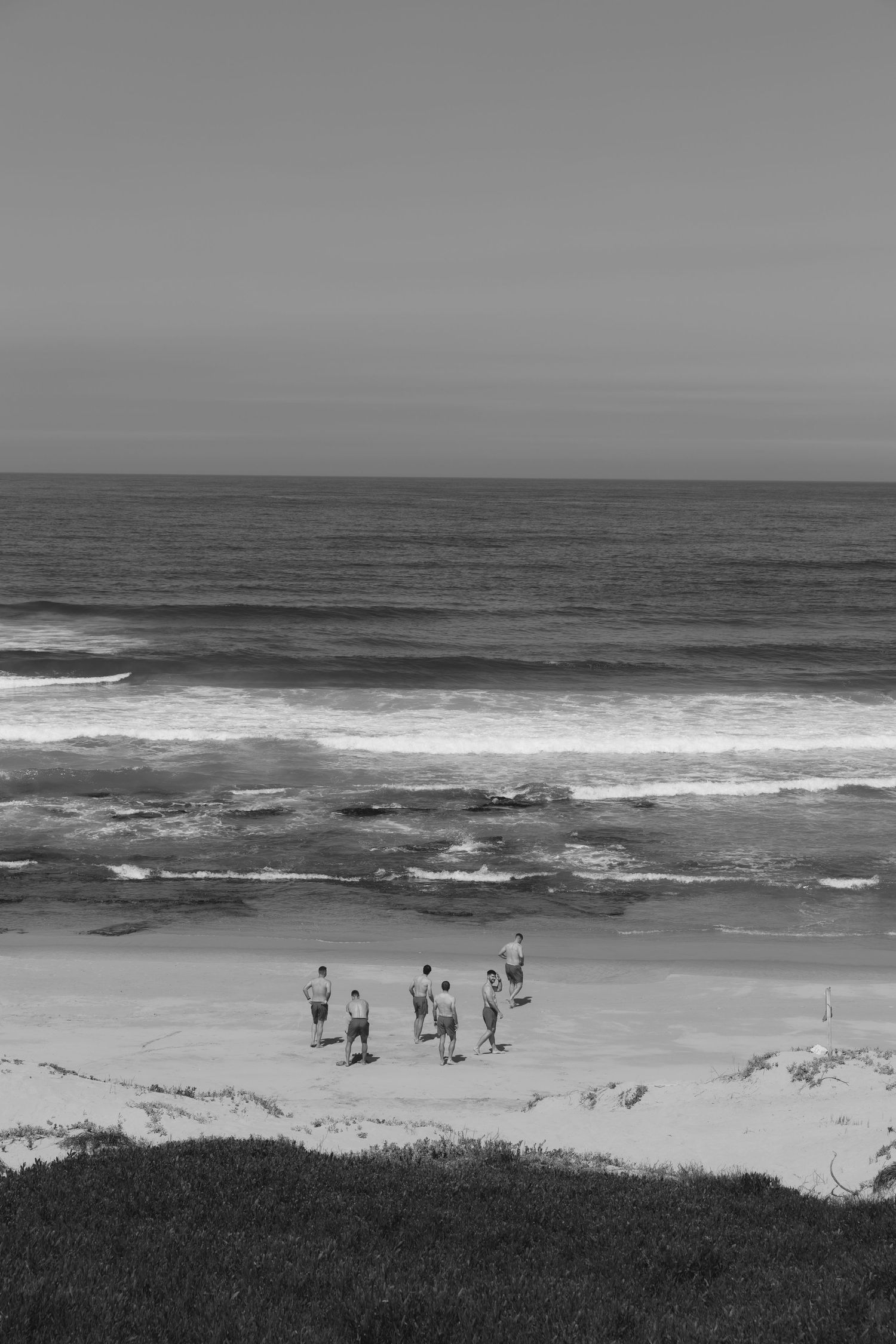 A panoramic black and white view of people walking along a coastal beach with crashing waves and grassy dunes.