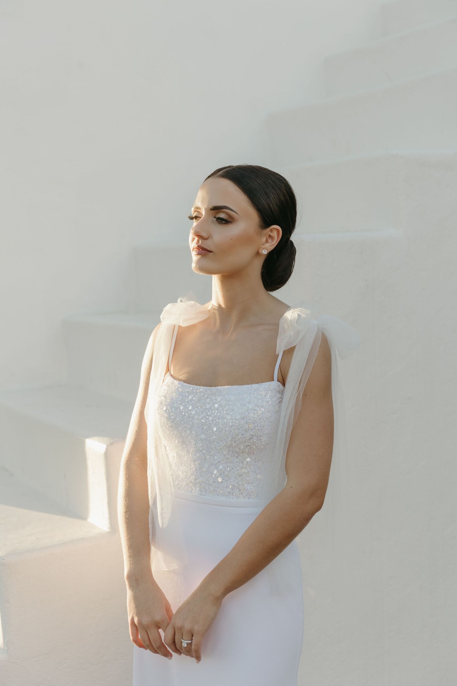 Elegant bride in a white beaded gown with sheer straps poses gracefully against a sunlit white wall.