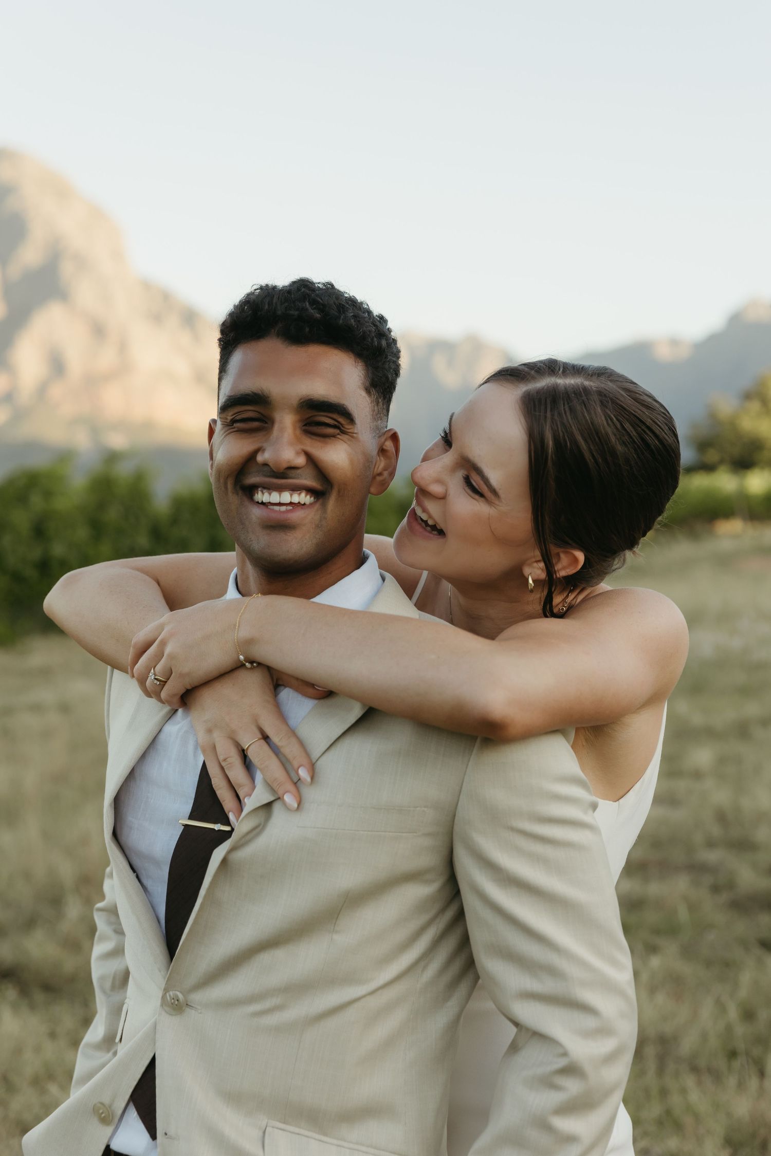Happy couple embracing outdoors in wedding attire with mountain backdrop, groom in beige suit, bride in white dress.