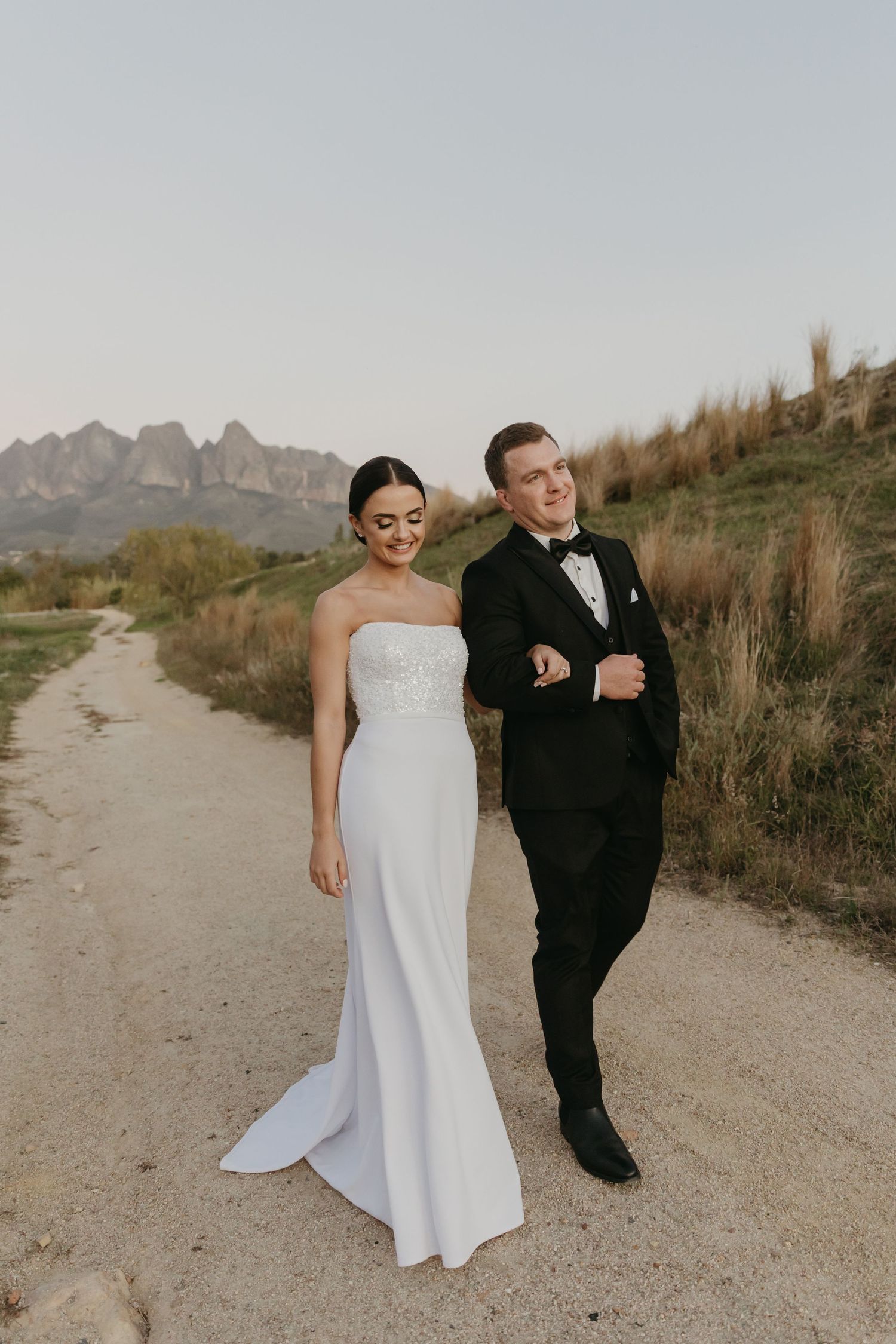Bride in white strapless gown and groom in black tuxedo walking on a dirt path with mountain scenery backdrop.
