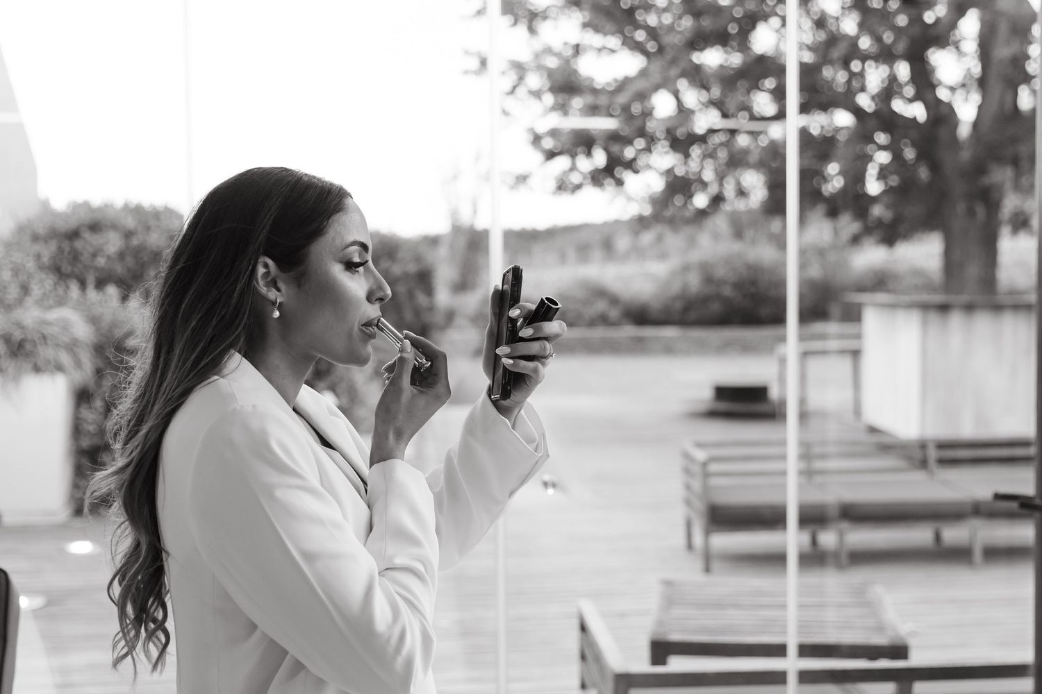 Black and white photo of a woman in a white blazer applying lipstick outdoors at an elegant venue.