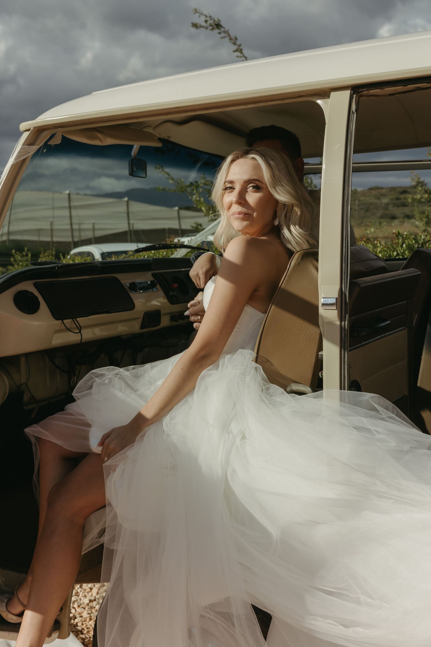 Bride in white tulle gown sitting in vintage van doorway, smiling with blonde hair, dramatic cloudy sky backdrop.