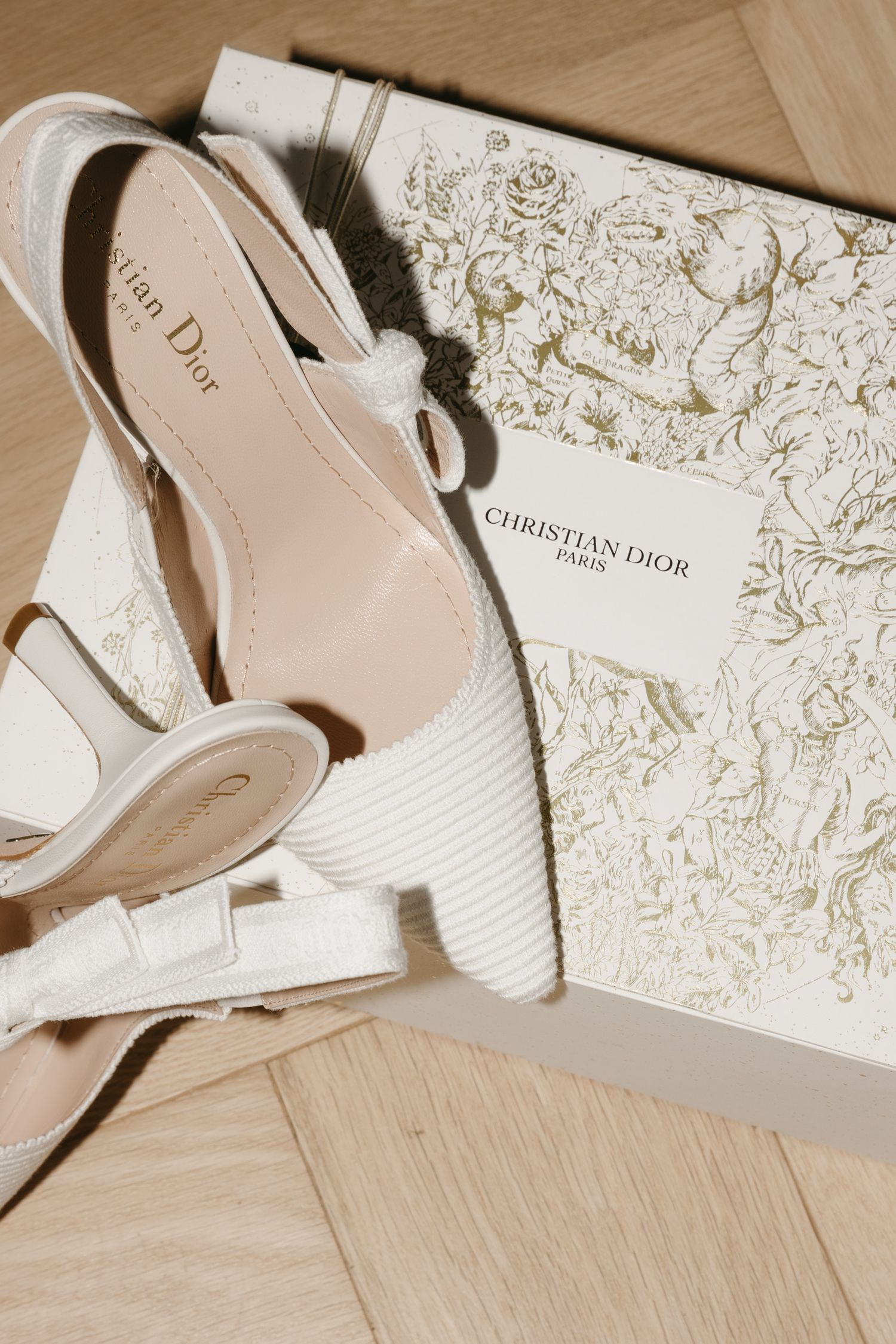 Christian Dior Paris white slingback heels with bow detail resting on branded luxury gift box on wooden floor.