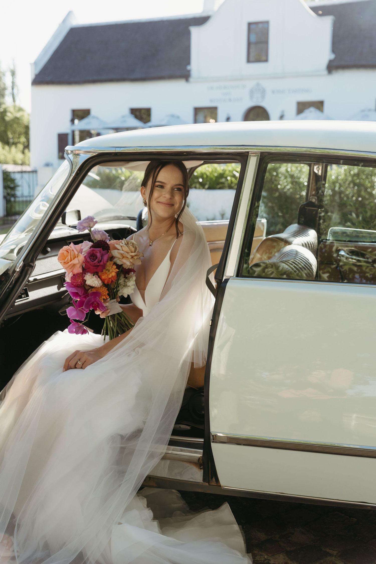Bride in white tulle gown holding colorful bouquet sitting in vintage car door at elegant white venue.