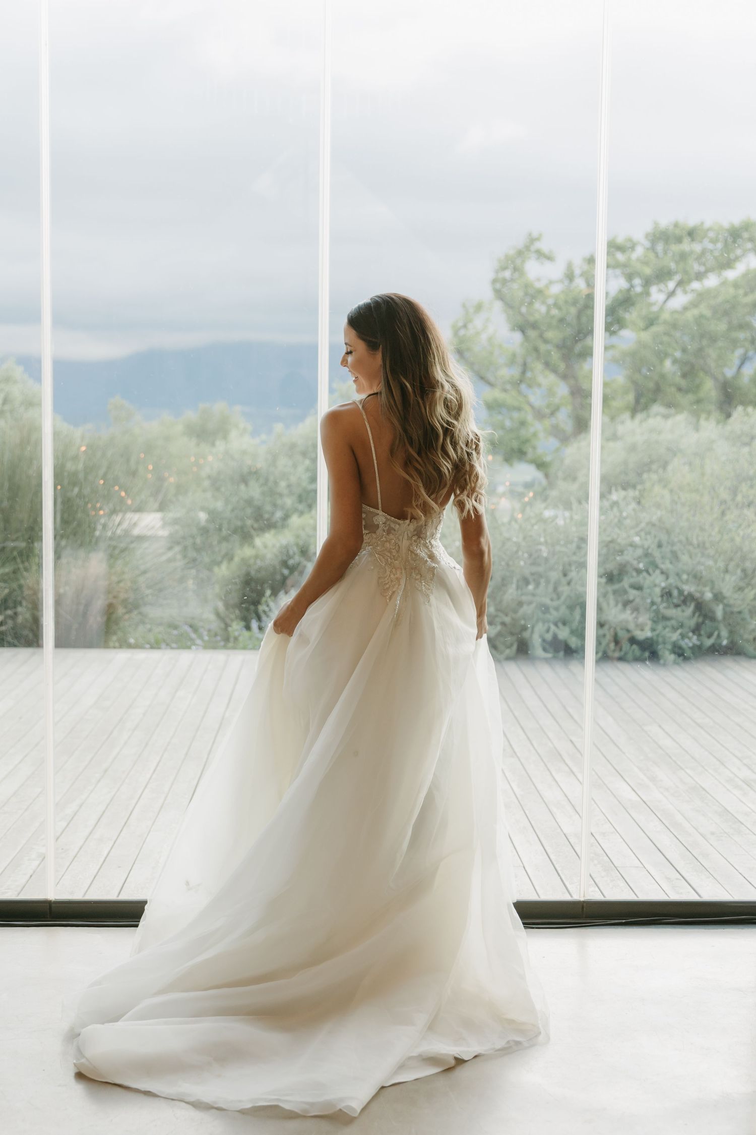 Bride in elegant backless white wedding gown with lace bodice stands before large windows with mountain views.