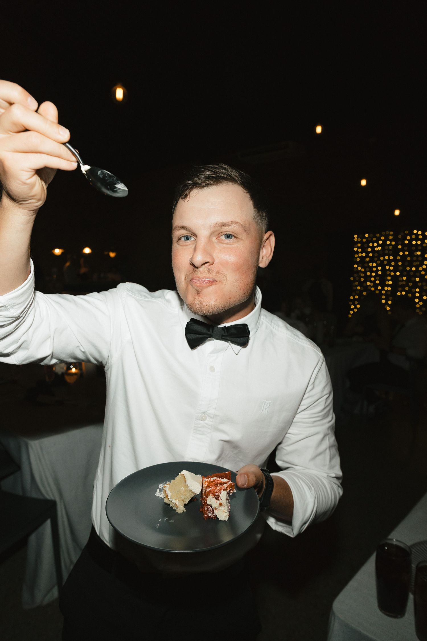 Person in white dress shirt and black bowtie eating food at a dimly lit event with string lights in background.