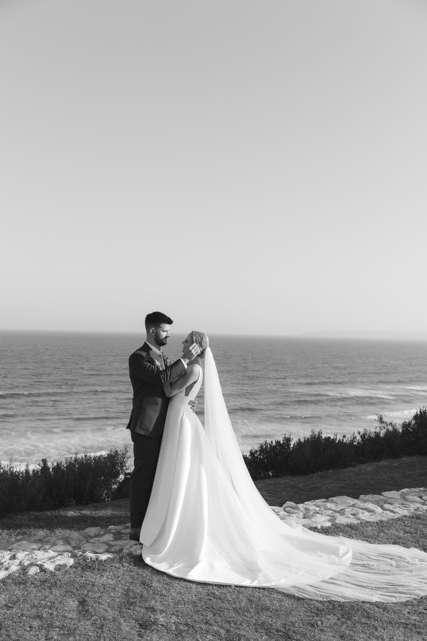 A black and white wedding photo series showing a couple posing against an ocean backdrop with a flowing white dress.