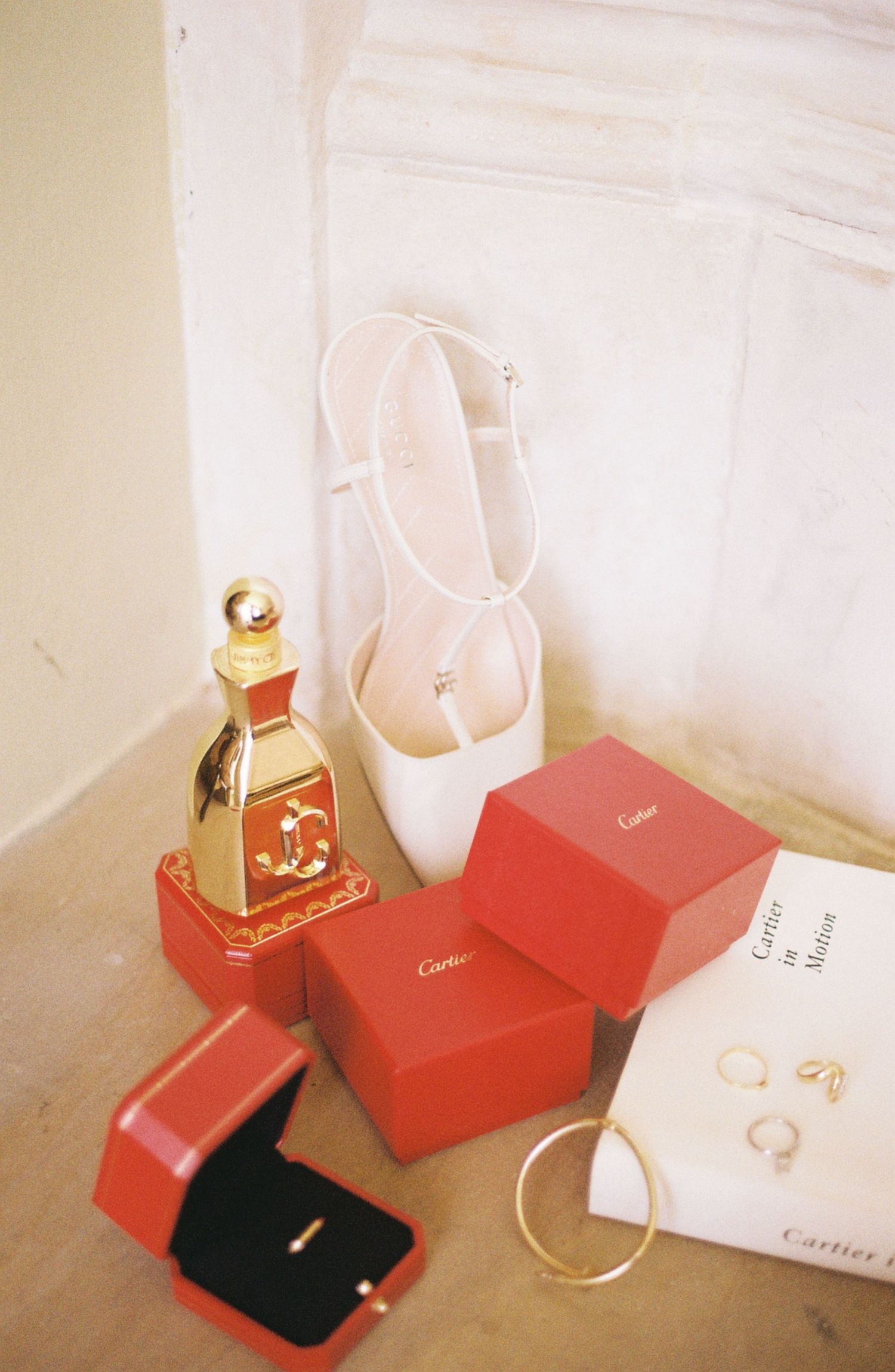 Elegant wedding accessories including white ballet shoes, Cartier red boxes, perfume bottle and jewelry on light surface.
