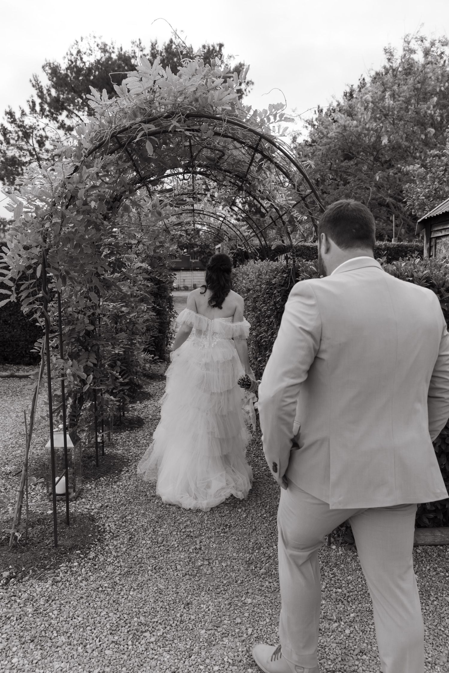 Black and white photo of bride and groom walking through a floral garden arch at their wedding.
