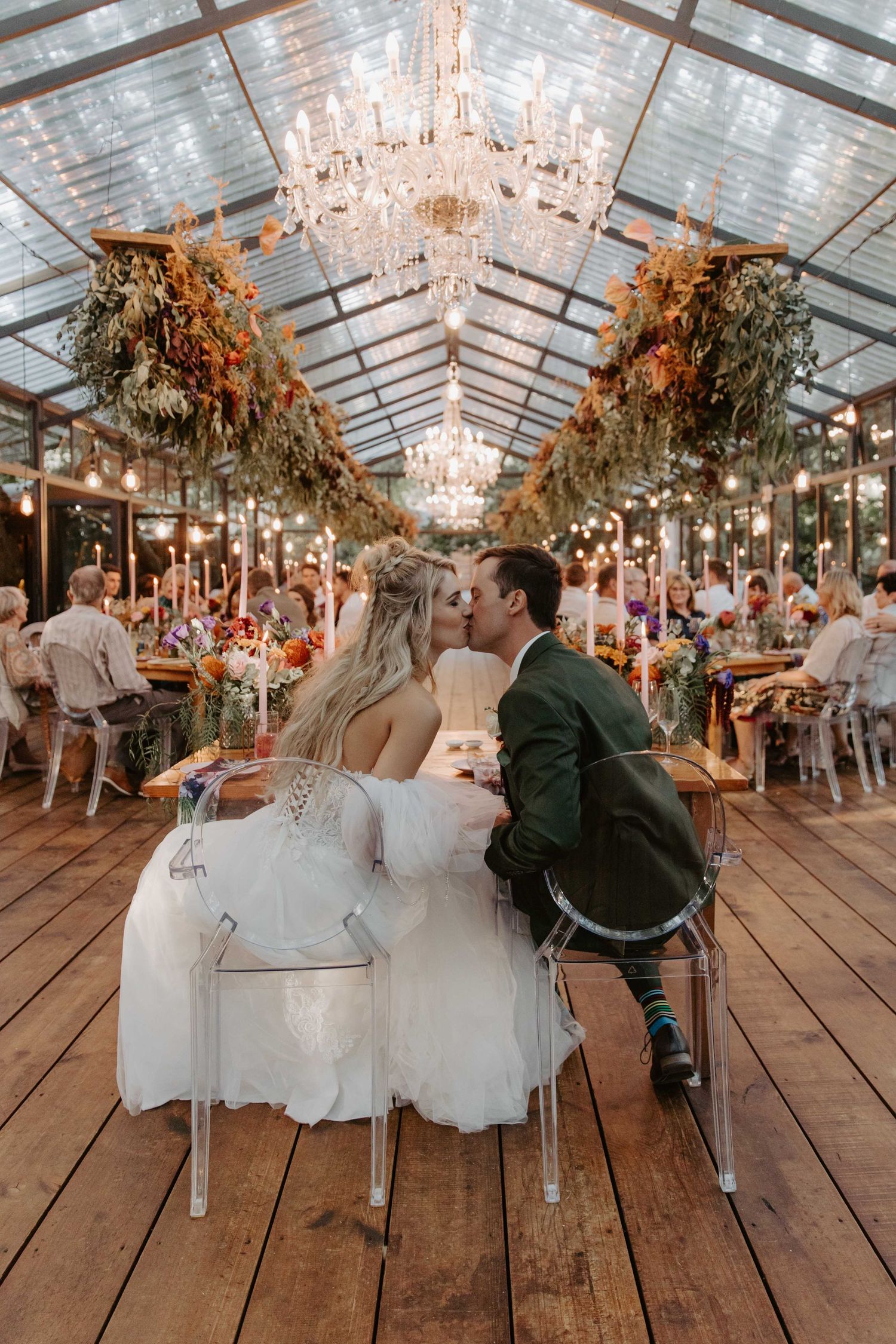 Bride and groom share a kiss at their elegant greenhouse wedding reception with chandeliers and colorful floral arrangements.