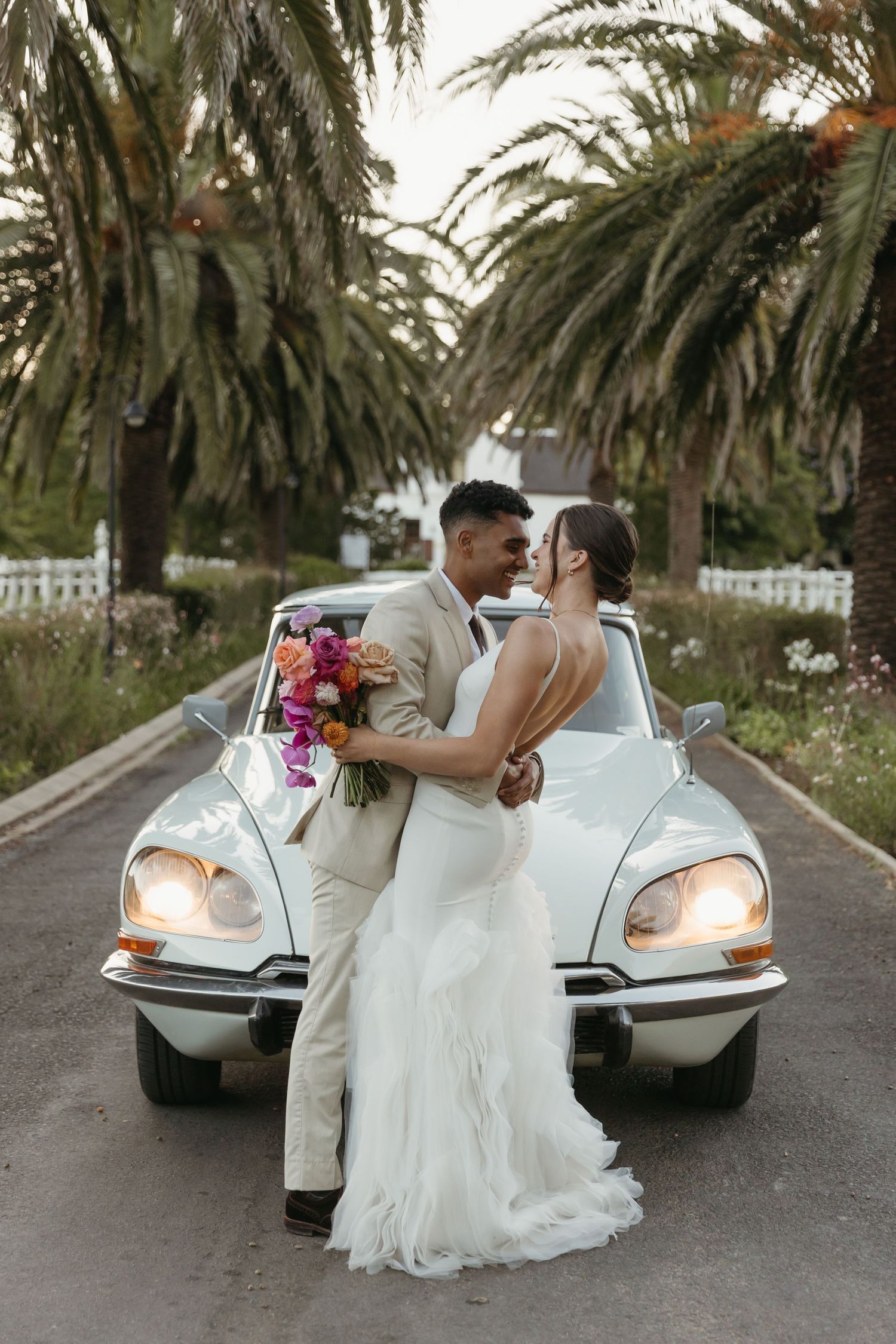 Newlywed couple embracing in front of a vintage white car, lined by palm trees, bride holding colorful bouquet.