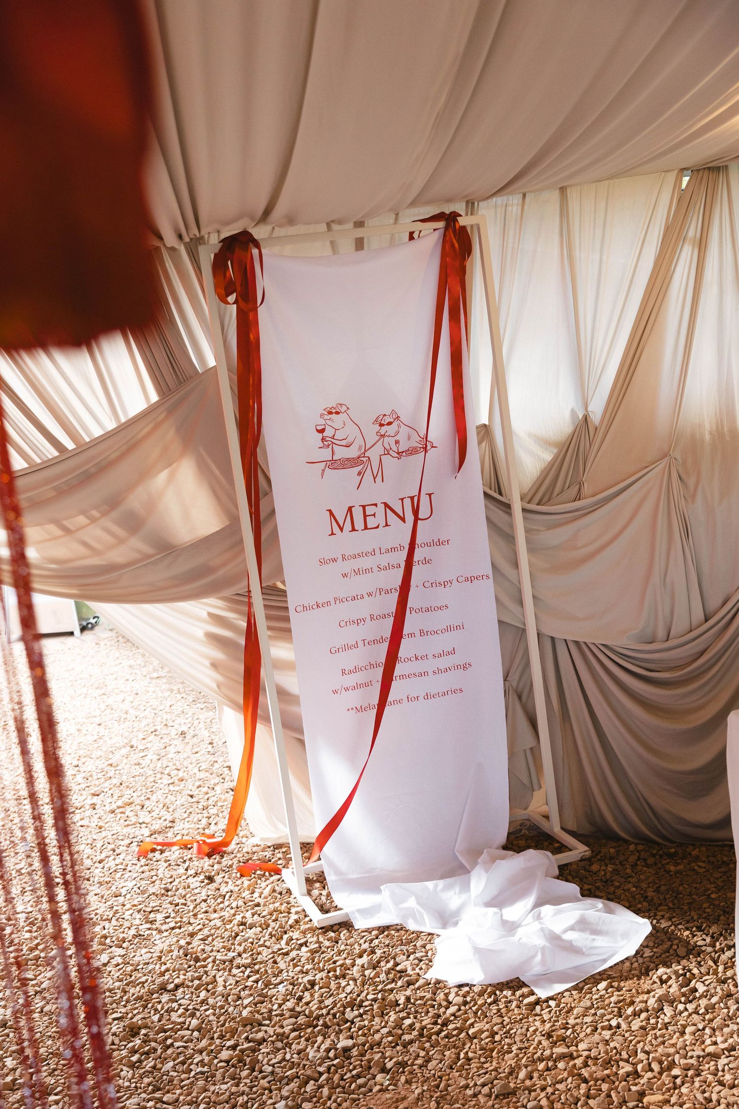 White fabric drapes and red ribbons create an elegant wedding tent atmosphere with menu signs and gravel flooring.