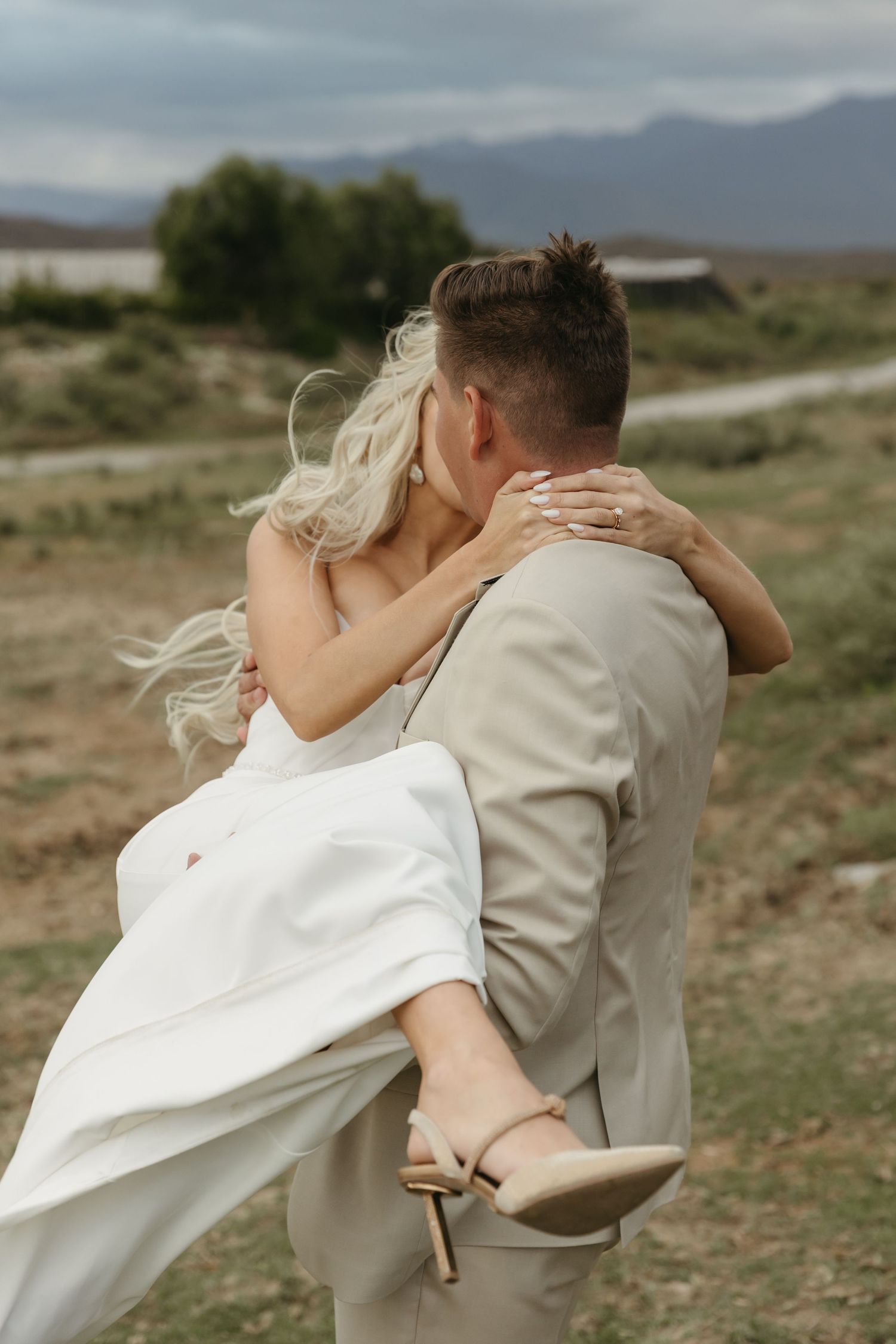 Groom in beige suit lifts bride in white wedding dress, sharing a romantic kiss outdoors with mountains in background.