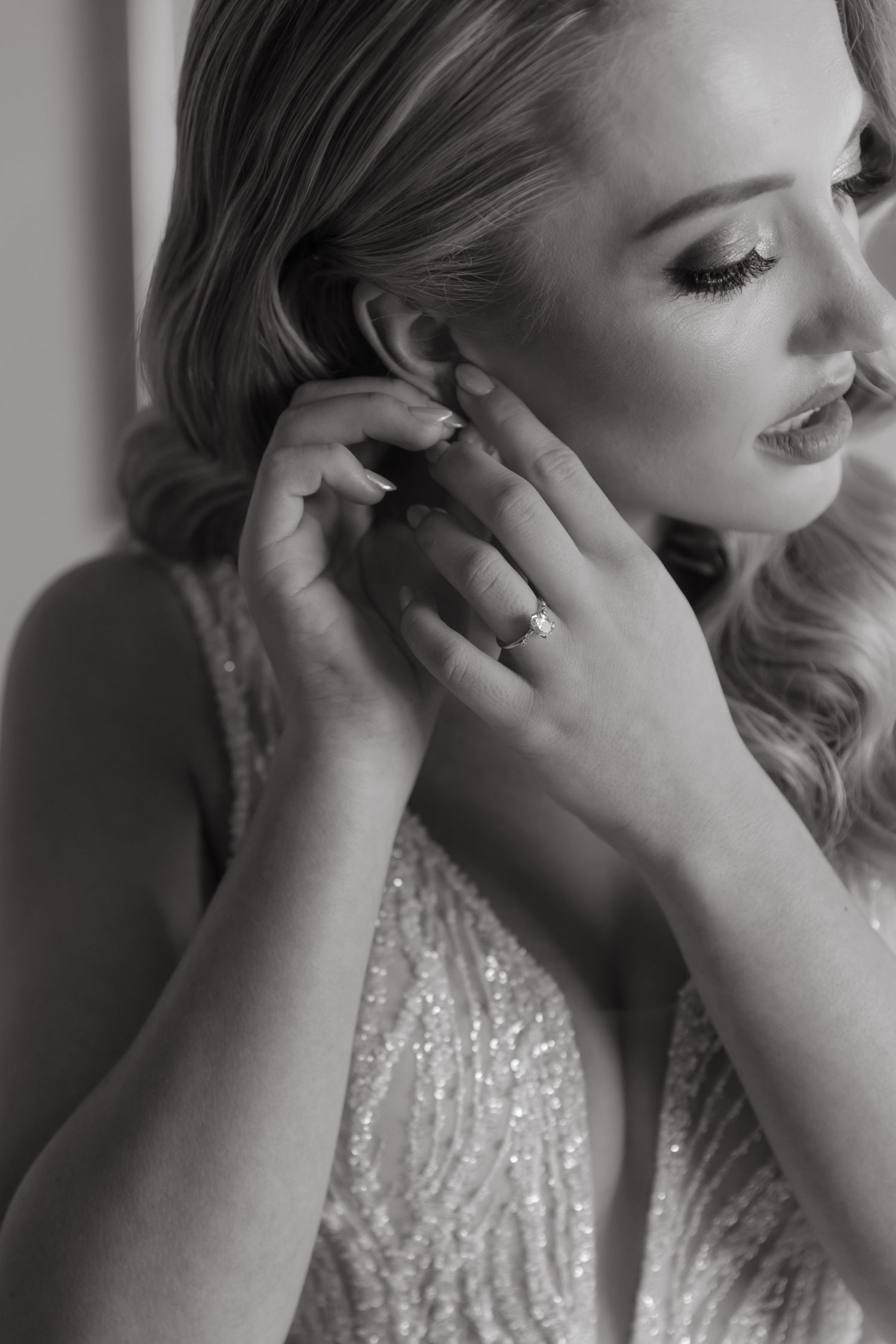 Black and white photo of elegant blonde bride putting on earring, wearing sparkly dress and diamond ring.