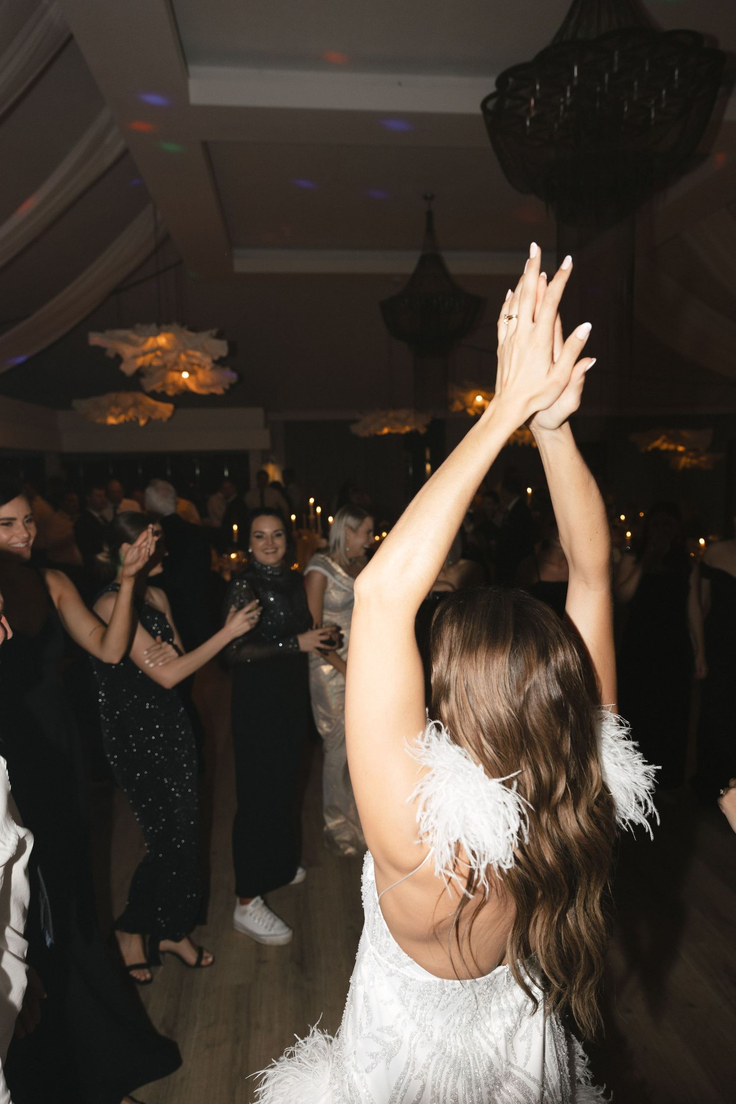 People dancing and celebrating at a party with raised hands and white feathered outfits in a dimly lit venue.
