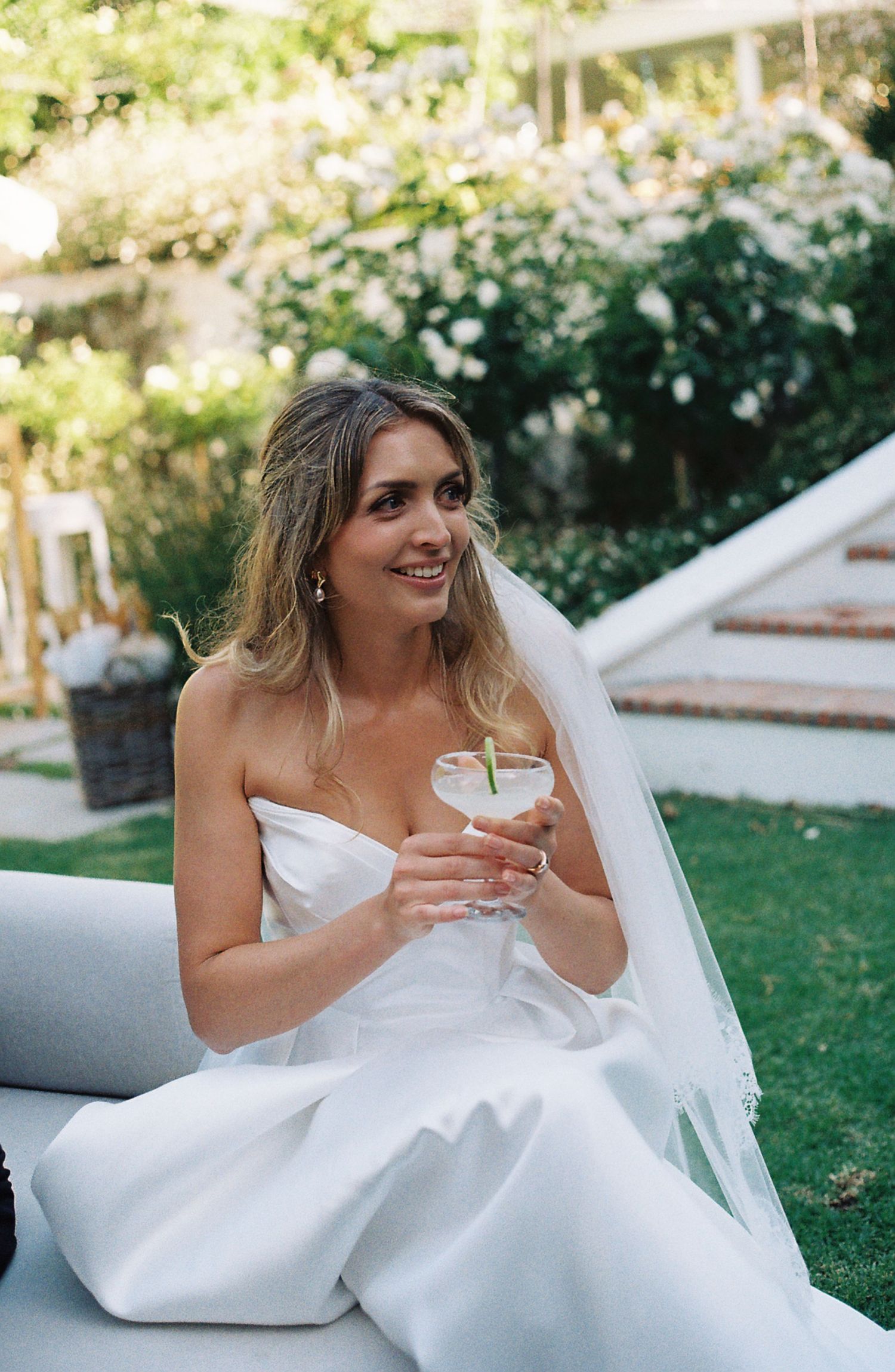 Bride in white wedding dress sits outdoors enjoying a drink during golden hour photography session.