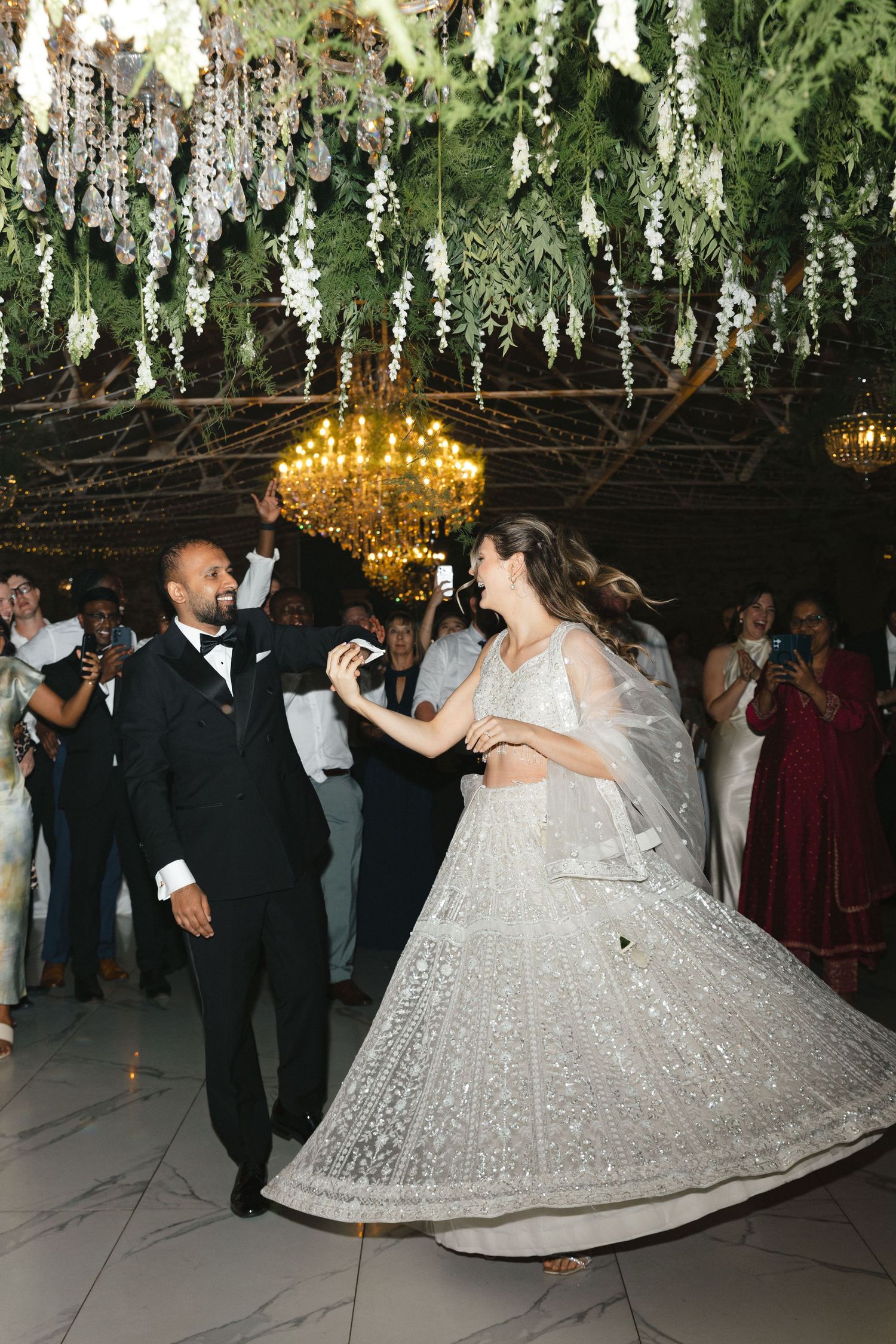 A couple dances together at an elegant wedding reception under hanging floral decorations and crystal chandeliers.